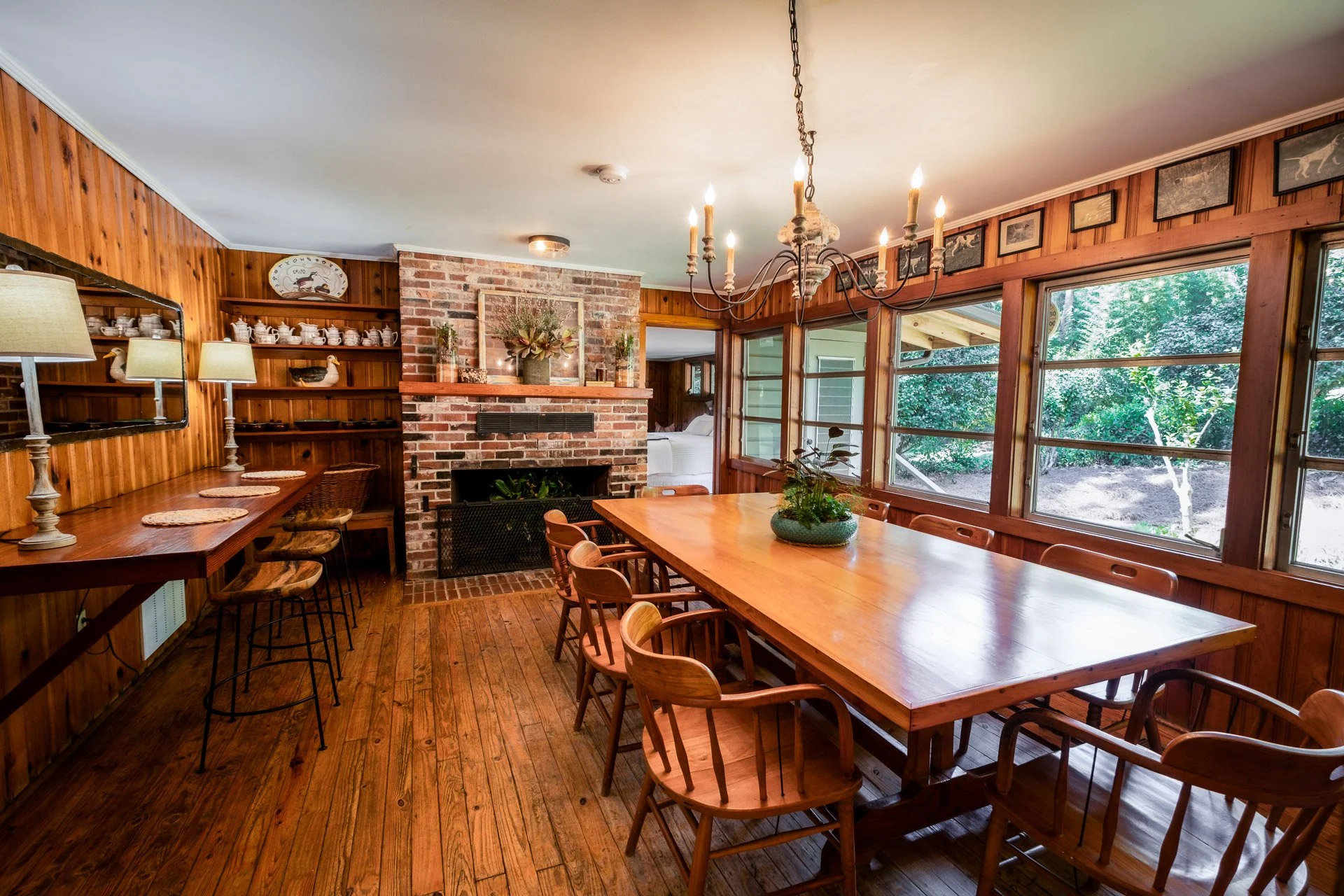 Wood-paneled dining room with a long wooden table, chairs, a brick fireplace, large windows showing green foliage outside, and a chandelier hanging from the ceiling.