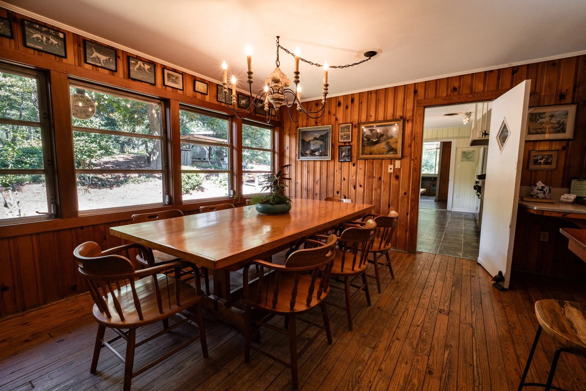 A rustic dining room with wooden walls and floor, large windows, a chandelier, and a wooden table with chairs. Artwork decorates the walls, and an open doorway leads to another room.