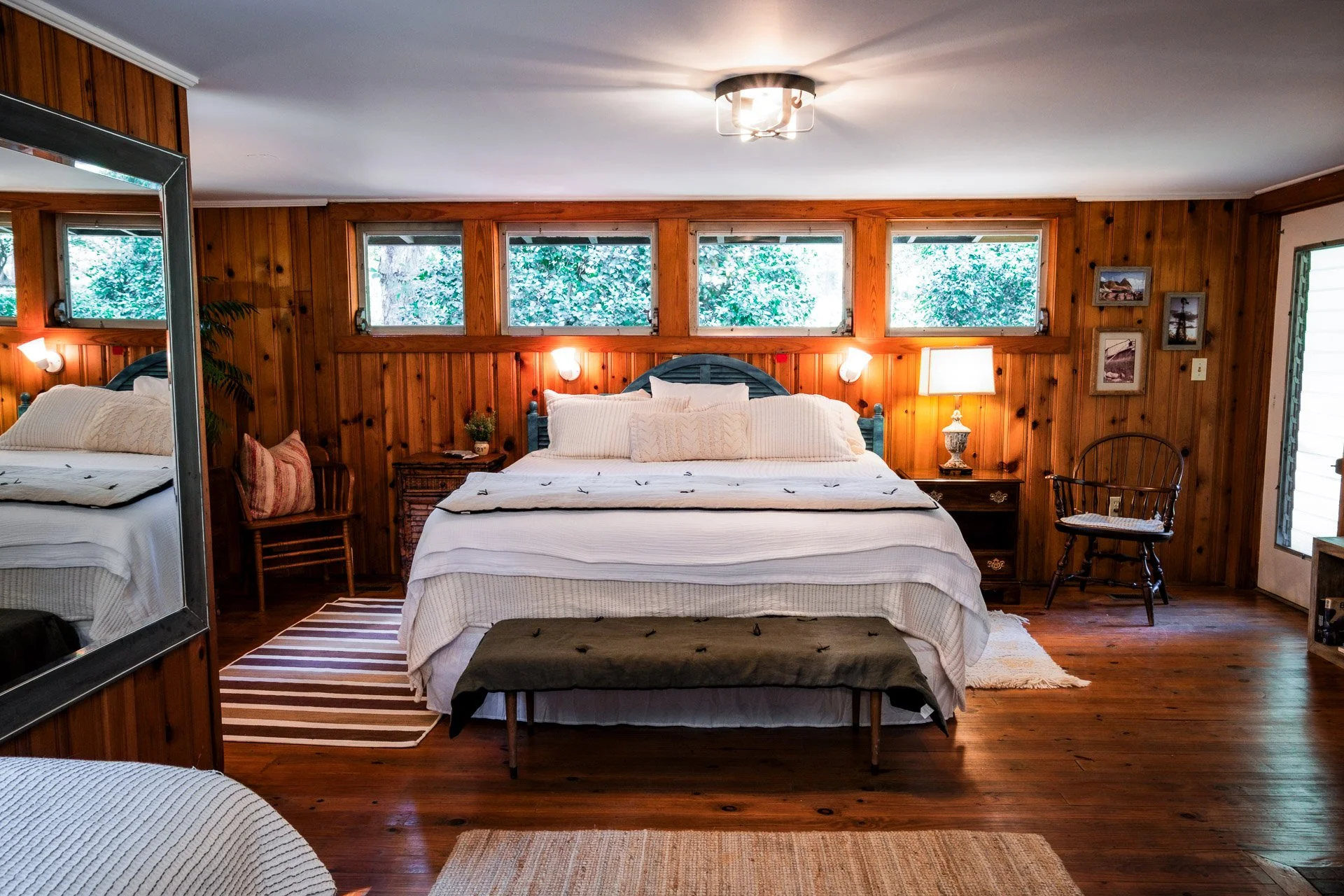 A cozy bedroom with wooden wall paneling, a large bed with white linens and two large pillows, bedside tables with lamps, a wooden chair, a mirror, and three rectangular windows above the bed showing greenery outside.