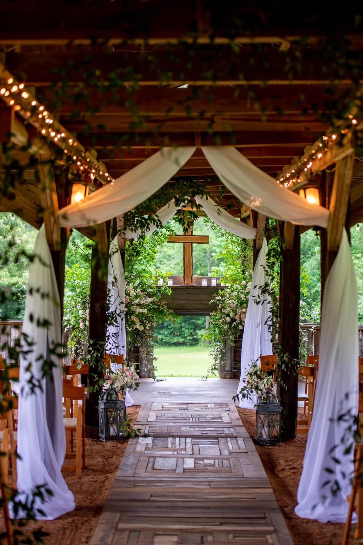 Decorated outdoor wedding aisle with white drapery, floral arrangements, lanterns, and a cross at the altar surrounded by greenery.