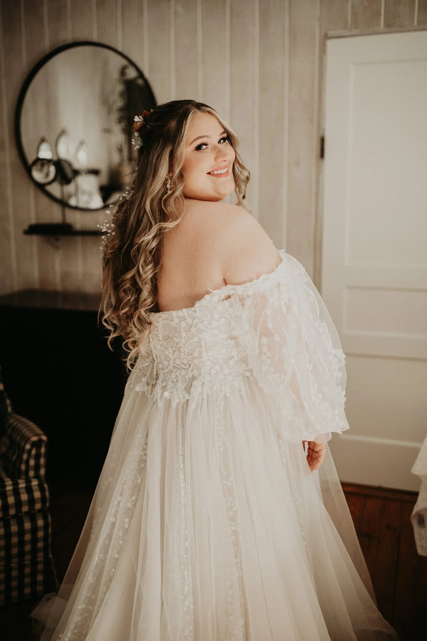 A woman in a white wedding dress with off-the-shoulder sleeves, smiling and looking over her shoulder.