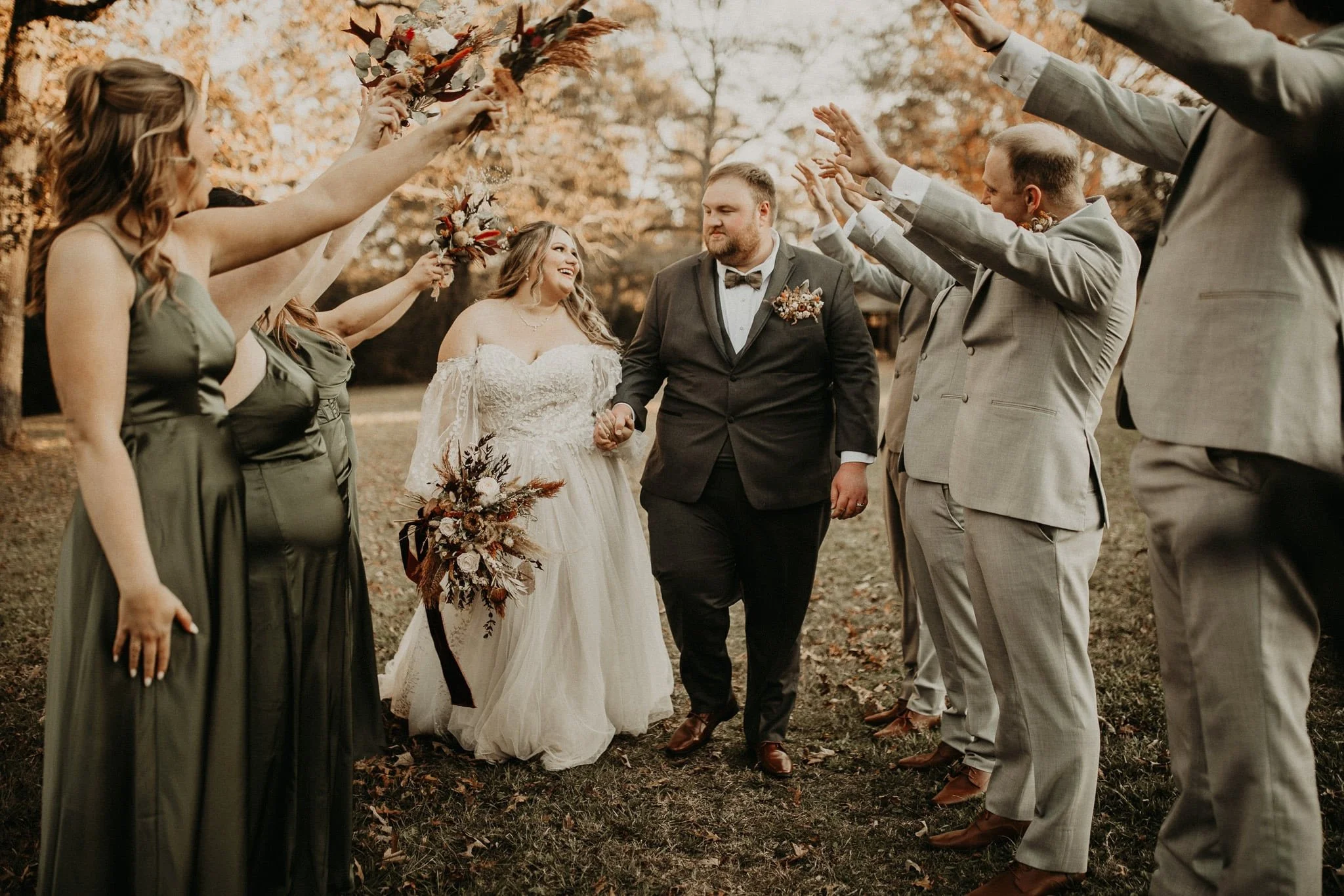 Bride and groom walking outdoors on their wedding day, holding hands, surrounded by bridesmaids in green dresses and groomsmen in light gray suits, with autumn trees and leaves in the background.