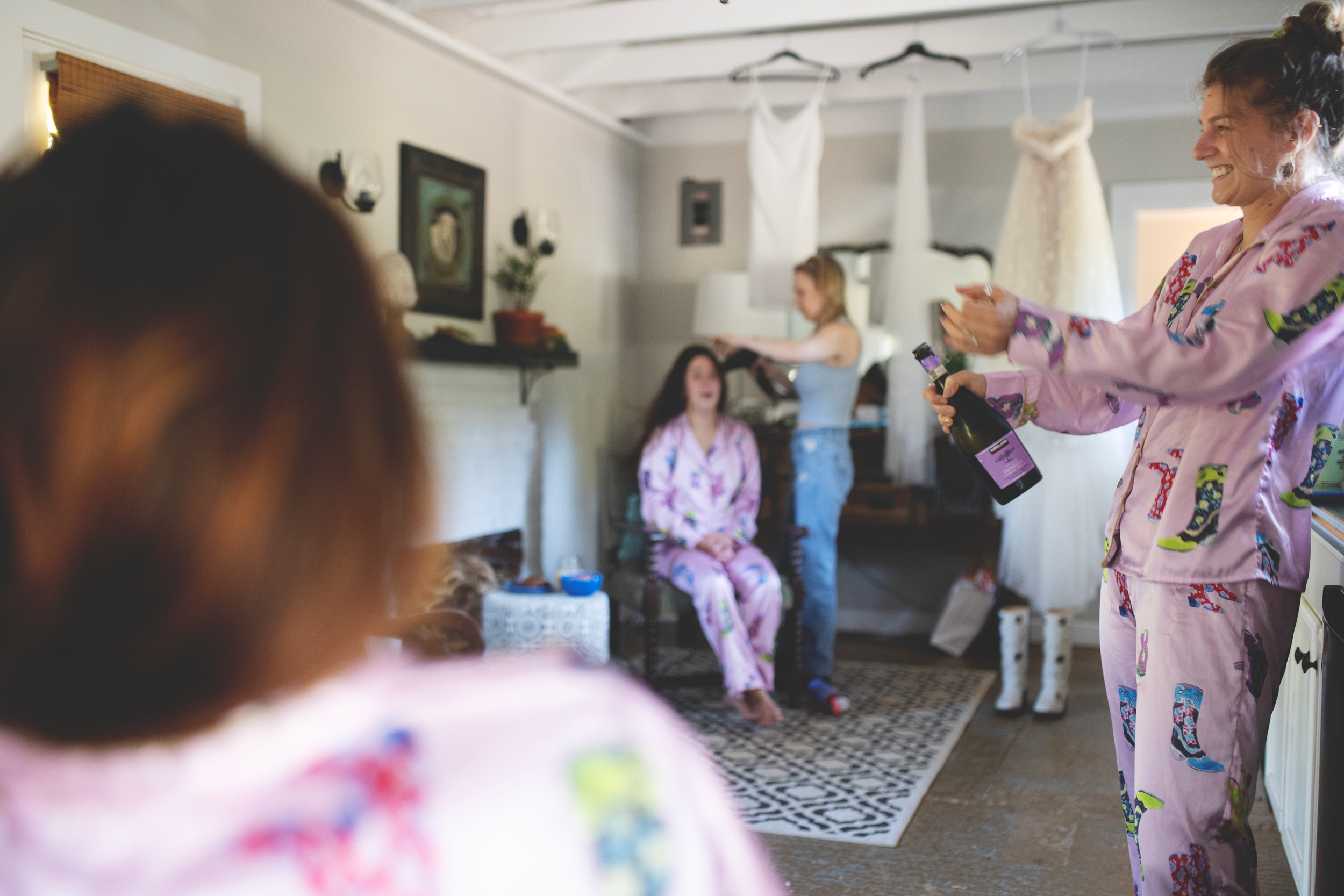 Women celebrating with champagne in a cozy living room, one woman in pink pajamas opening a bottle, others in the background preparing for celebration.