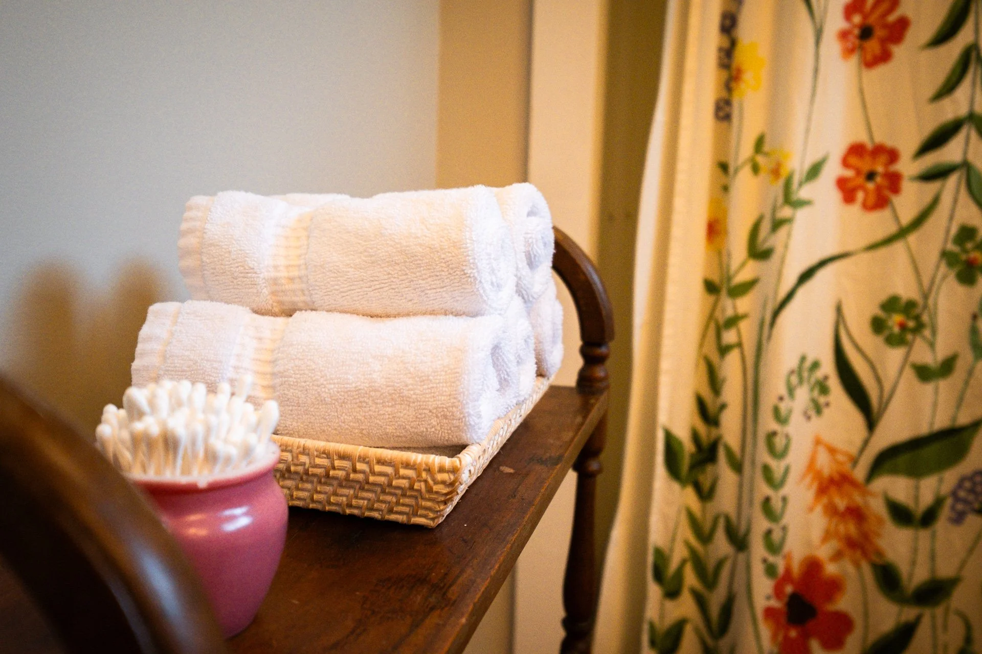 Stacked white rolled towels on a wicker tray on a wooden table, with a pink container filled with cotton swabs nearby and floral patterned curtains in the background.