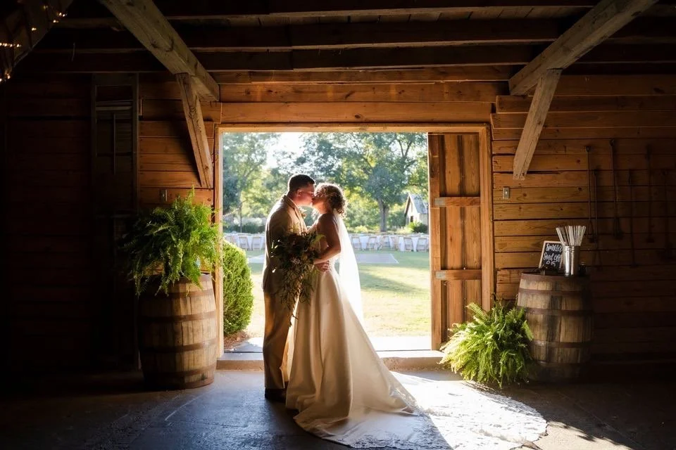 A bride and groom standing inside a wooden barn, sharing a kiss as sunlight shines around them, with trees and chairs visible outside.
