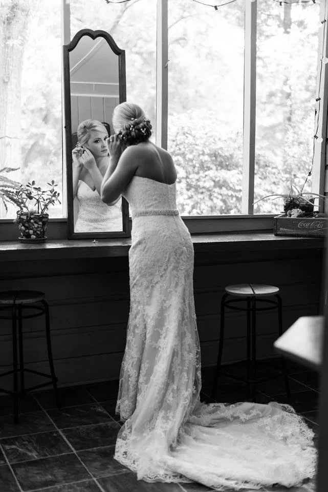 A woman in a wedding dress looking at herself in a mirror on a window sill.