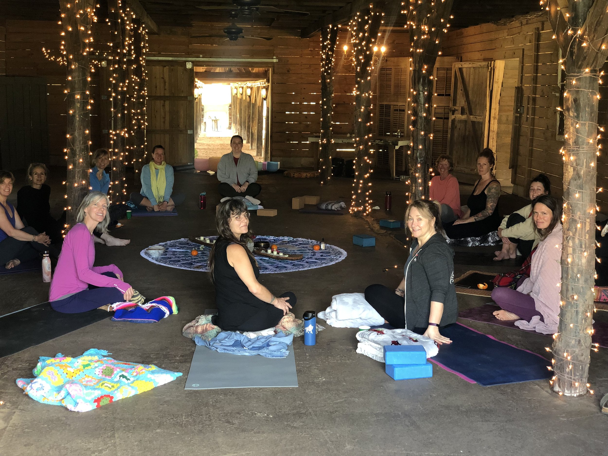 Group of people participating in a yoga or meditation session in a rustic wooden barn, sitting on mats with string lights decorating the pillars and ceiling, with open barn doors revealing the outside.