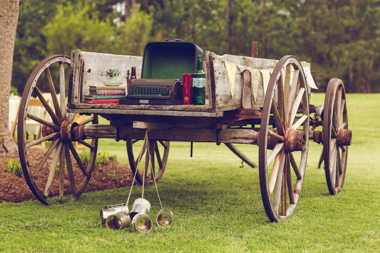 An old wooden wagon with large spoked wheels, used as a table, set outdoors in a grassy area with trees. On top of the wagon are books, a vintage typewriter, a green bottle, a small bowl with a plant, and an open green suitcase. Several empty cans are hanging from the wagon's side and lying on the grass.