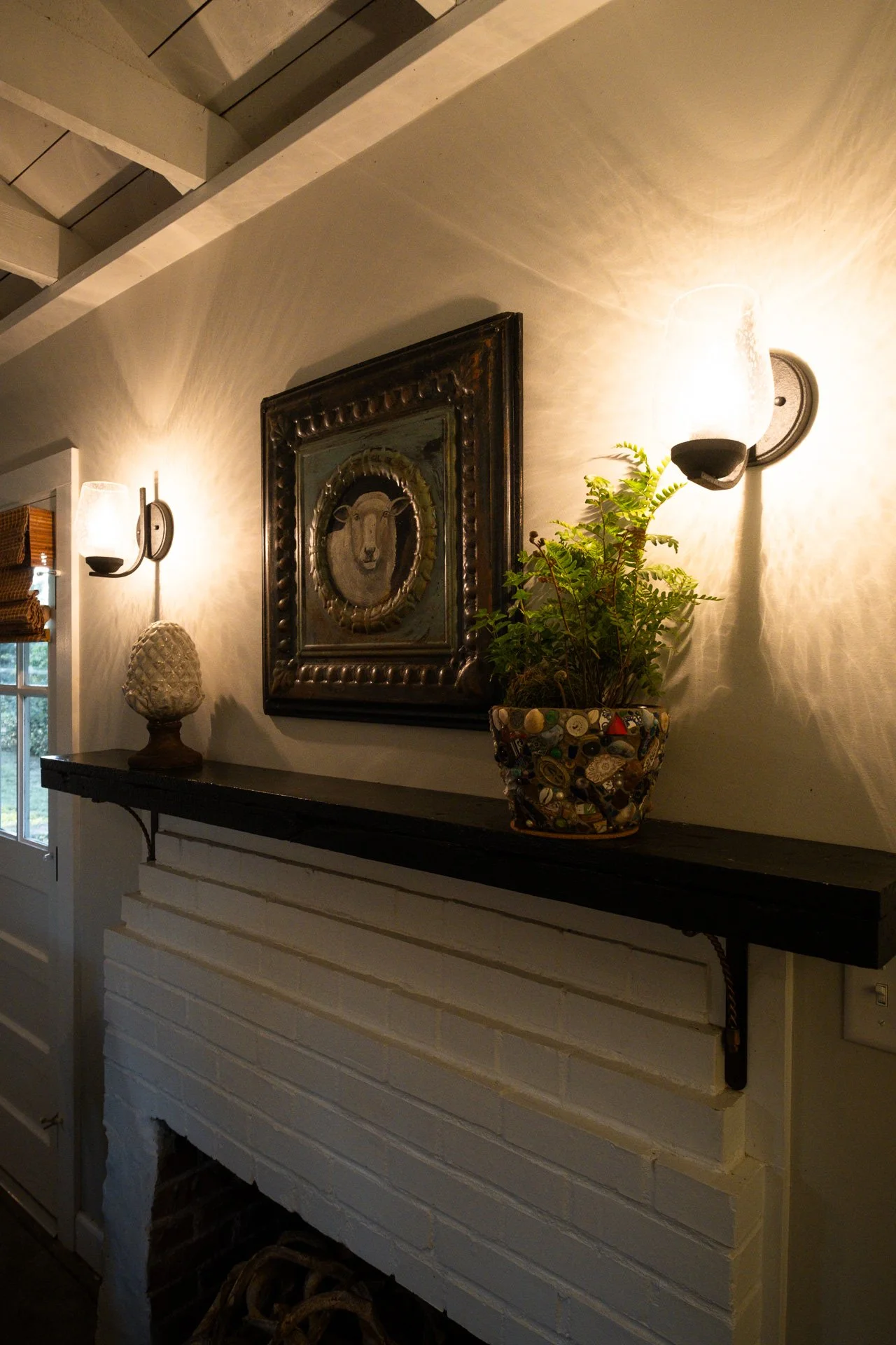 A cozy living room fireplace with a white painted brick surround, a black mantel, and decorative decor including a framed art piece, a potted plant, a sculptural lamp, and wall-mounted sconces that cast warm light, creating a welcoming ambiance.