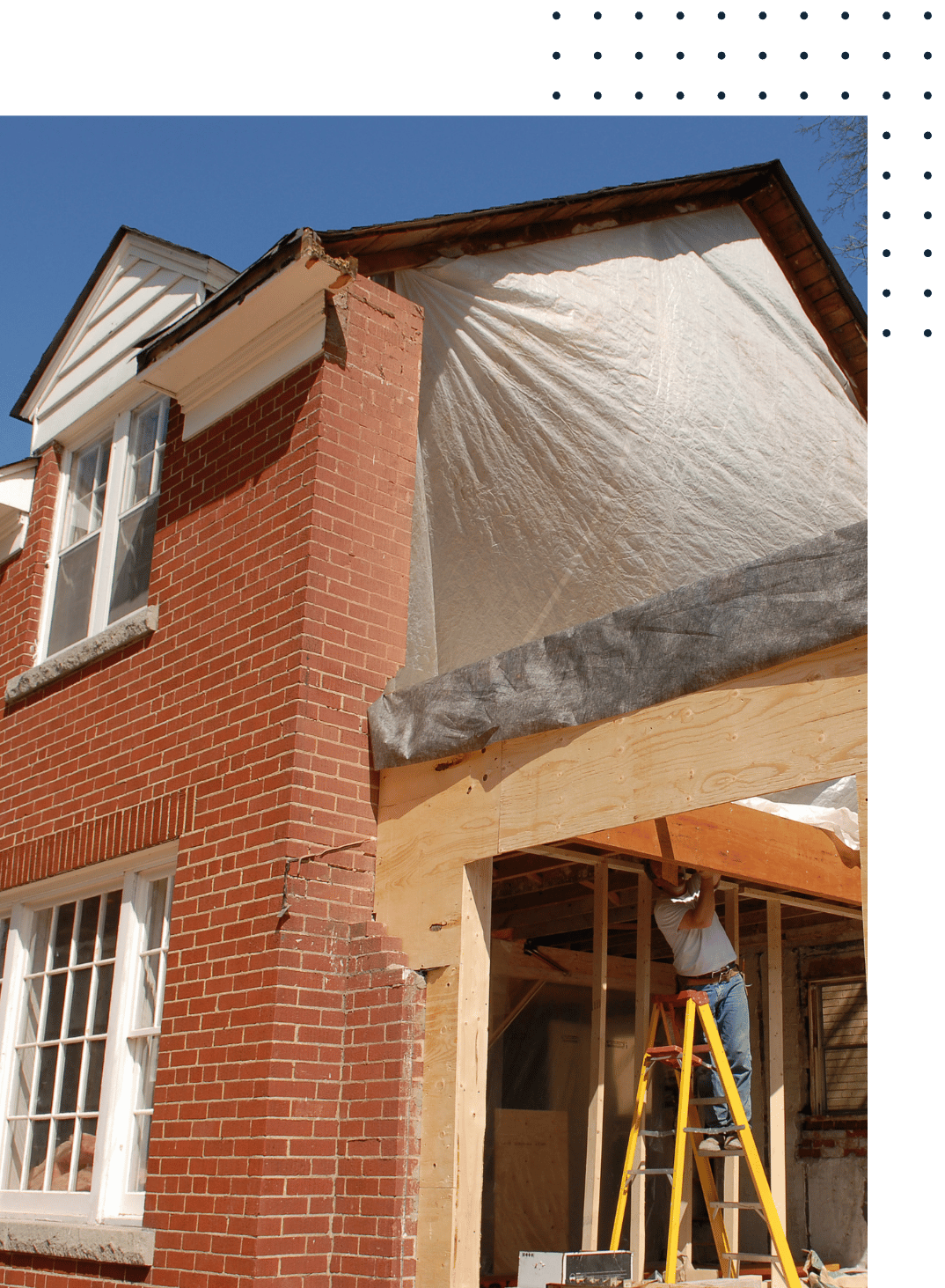Construction worker on a ladder working on the wooden frame of an extension or renovation to a brick house under a clear blue sky.