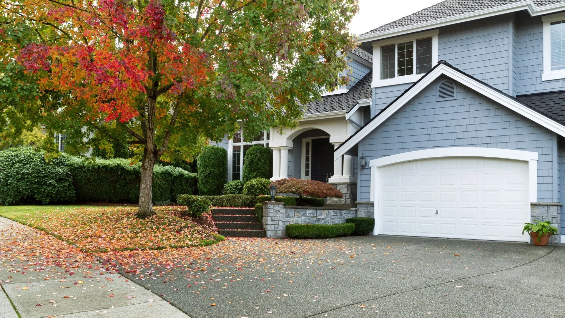 A two-story blue house with white trim and a white garage door, surrounded by green shrubs and trees with fall foliage, and a sidewalk with fallen leaves.