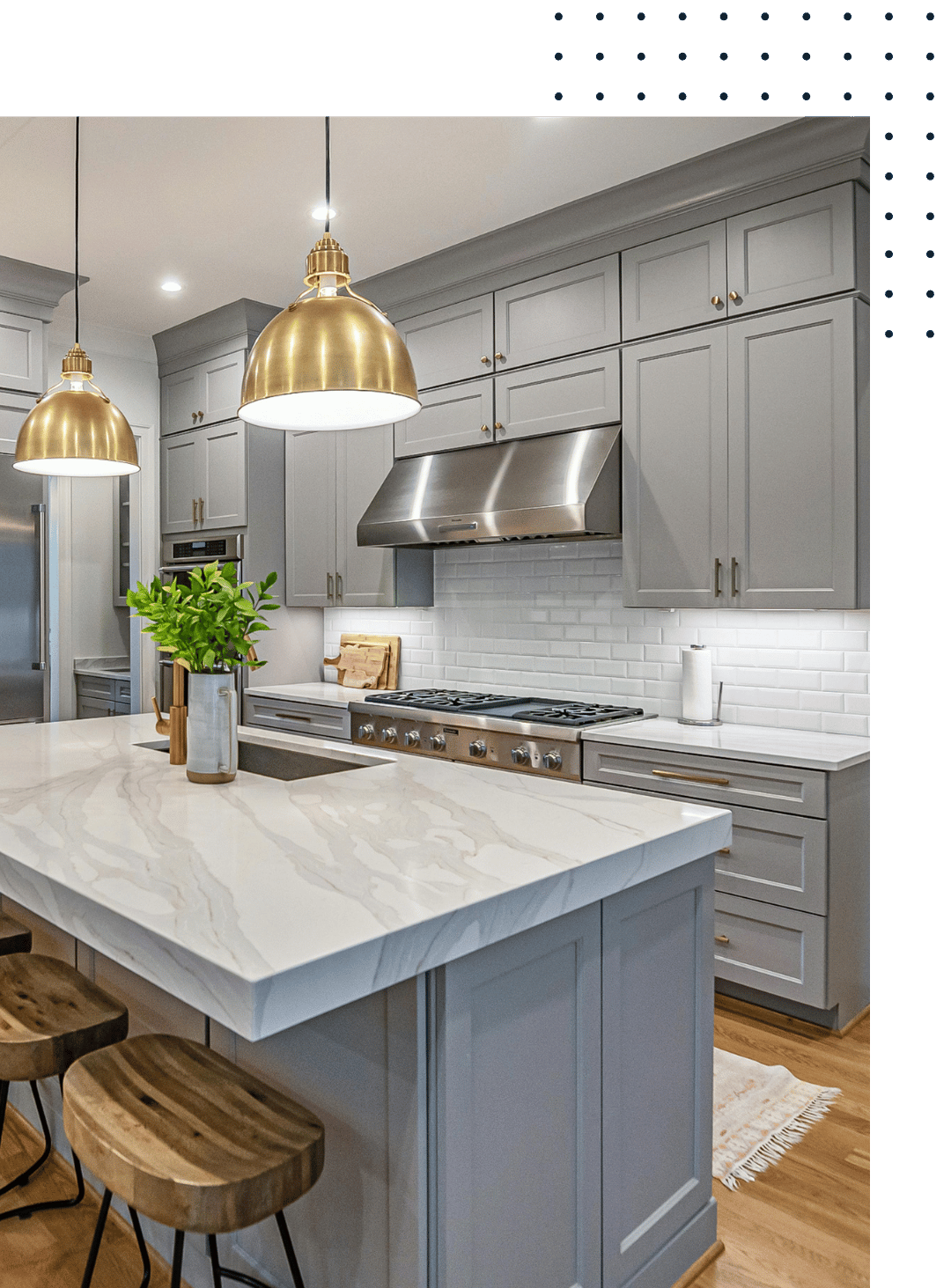 Modern kitchen with light gray cabinets, white marble countertop island, white subway tile backsplash, stainless steel appliances, and two gold pendant lights hanging over the island. There is a potted plant and wooden stools at the island, with hardwood floors and a white rug on the floor.