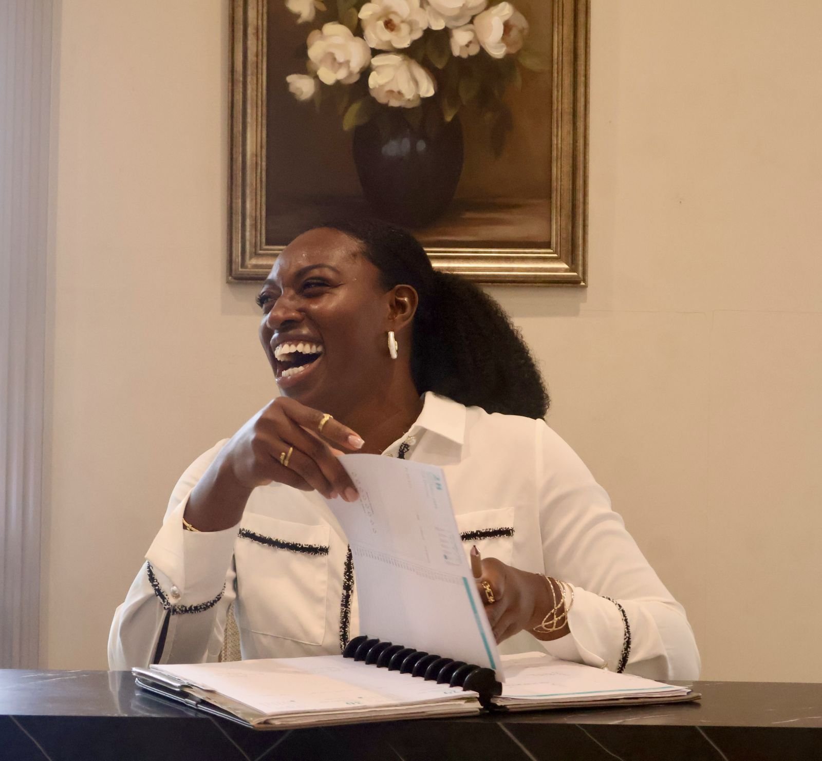 A woman sitting at a desk, smiling and laughing, holding a calendar, with a painting of white flowers in a black vase hanging on the wall behind her.