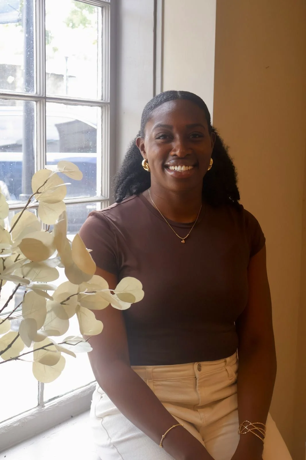 A woman with dark hair, gold jewelry, and a brown top sitting by a window with beige walls and white flower decoration.