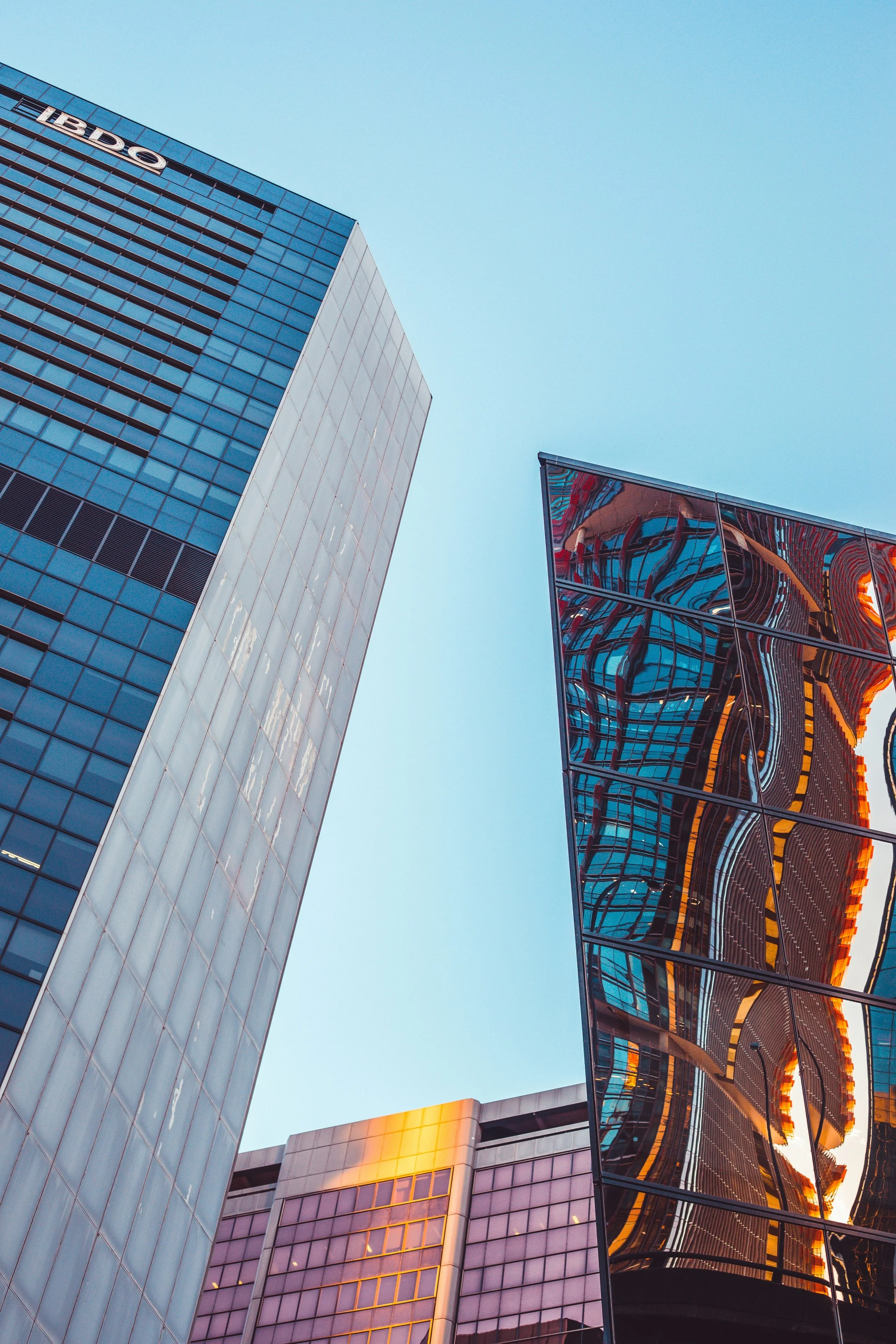 Several tall modern skyscrapers with reflective glass windows, one with a reflective surface showing distorted reflections of other buildings, against a clear blue sky.