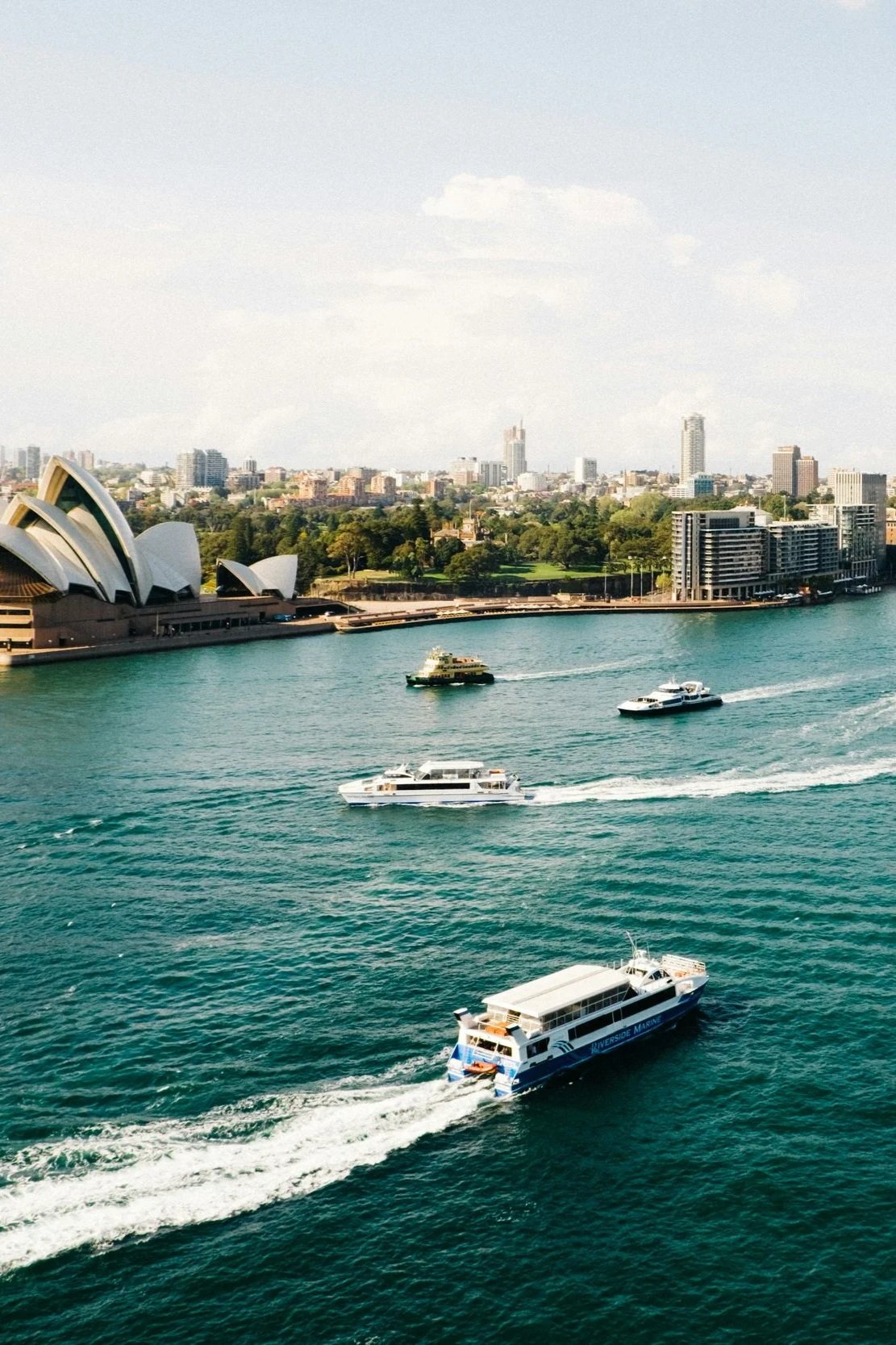 Multiple boats sailing on a waterway near the Sydney Opera House and city skyline.