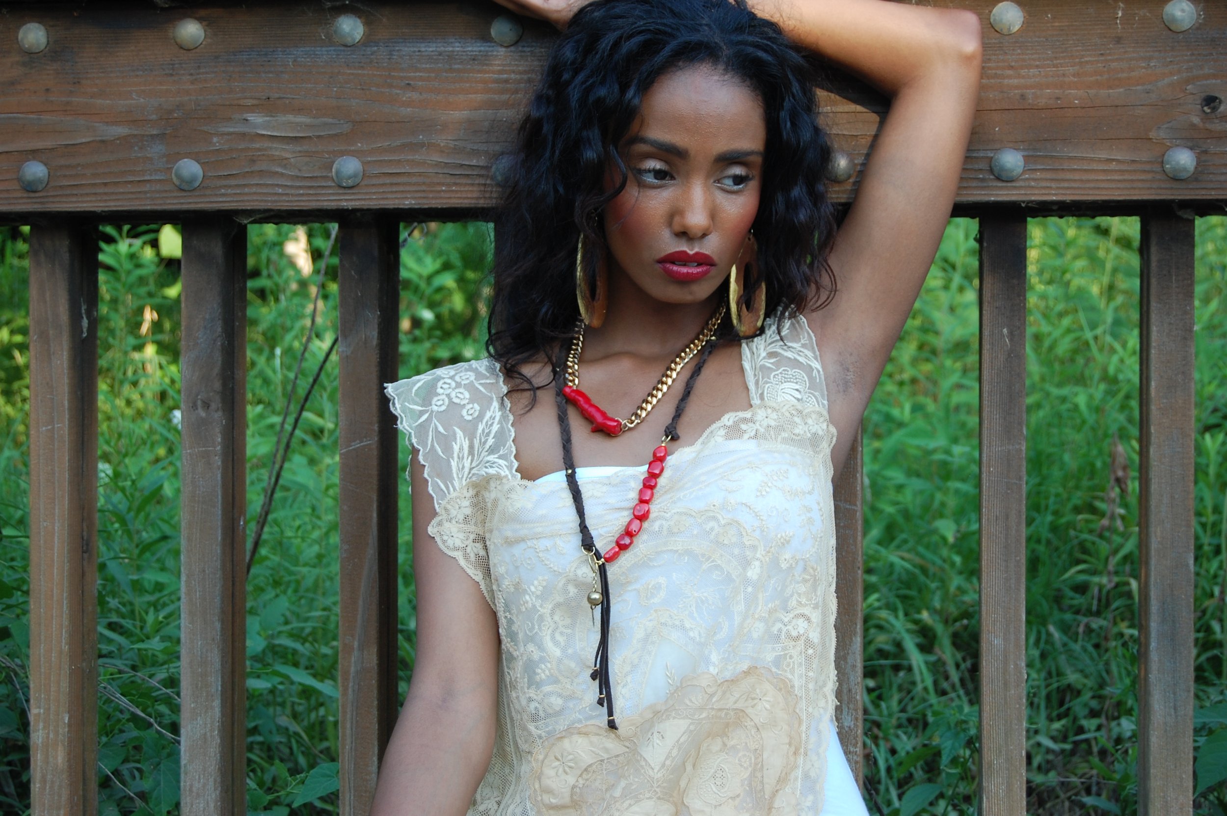 A woman with dark, curly hair, wearing a cream-colored lace dress and multiple necklaces, standing outdoors near a wooden fence with greenery in the background.
