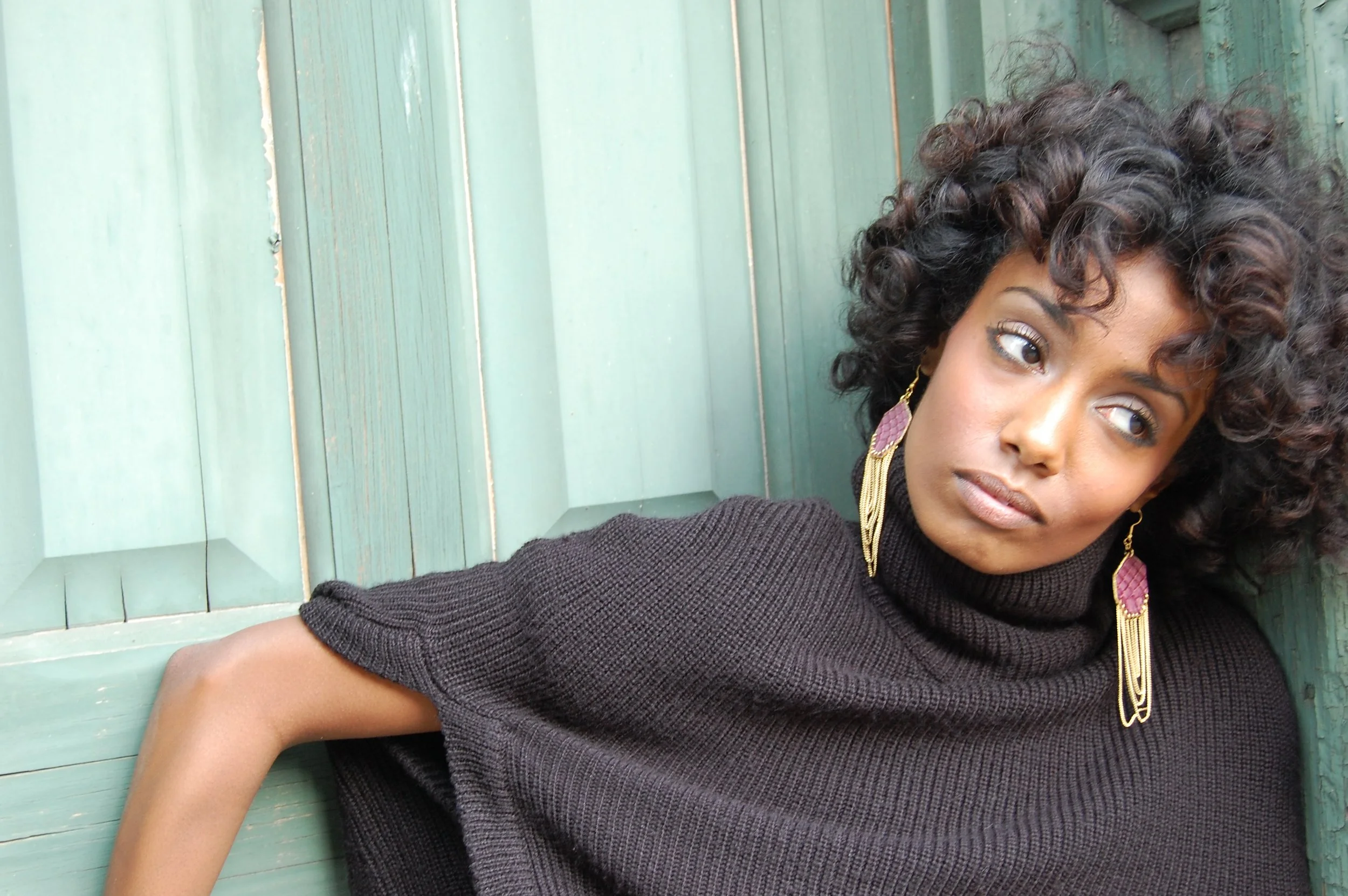 A woman with curly hair wearing a black turtleneck sweater and pink earrings, leaning against a green wooden wall.