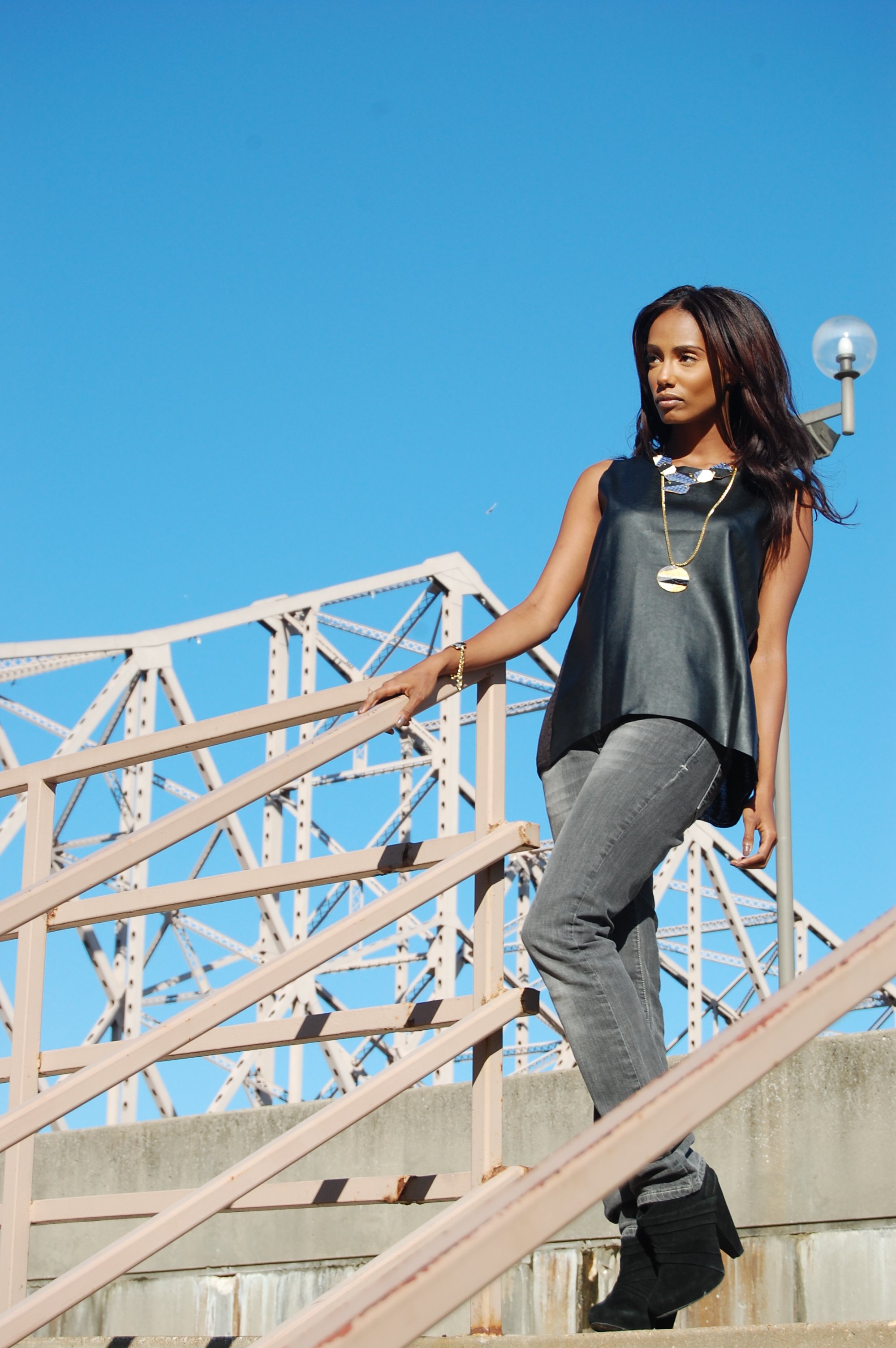 A woman with dark hair wearing a black sleeveless top, gray jeans, and black boots walking down outdoor stairs against a bright blue sky with a steel bridge in the background.
