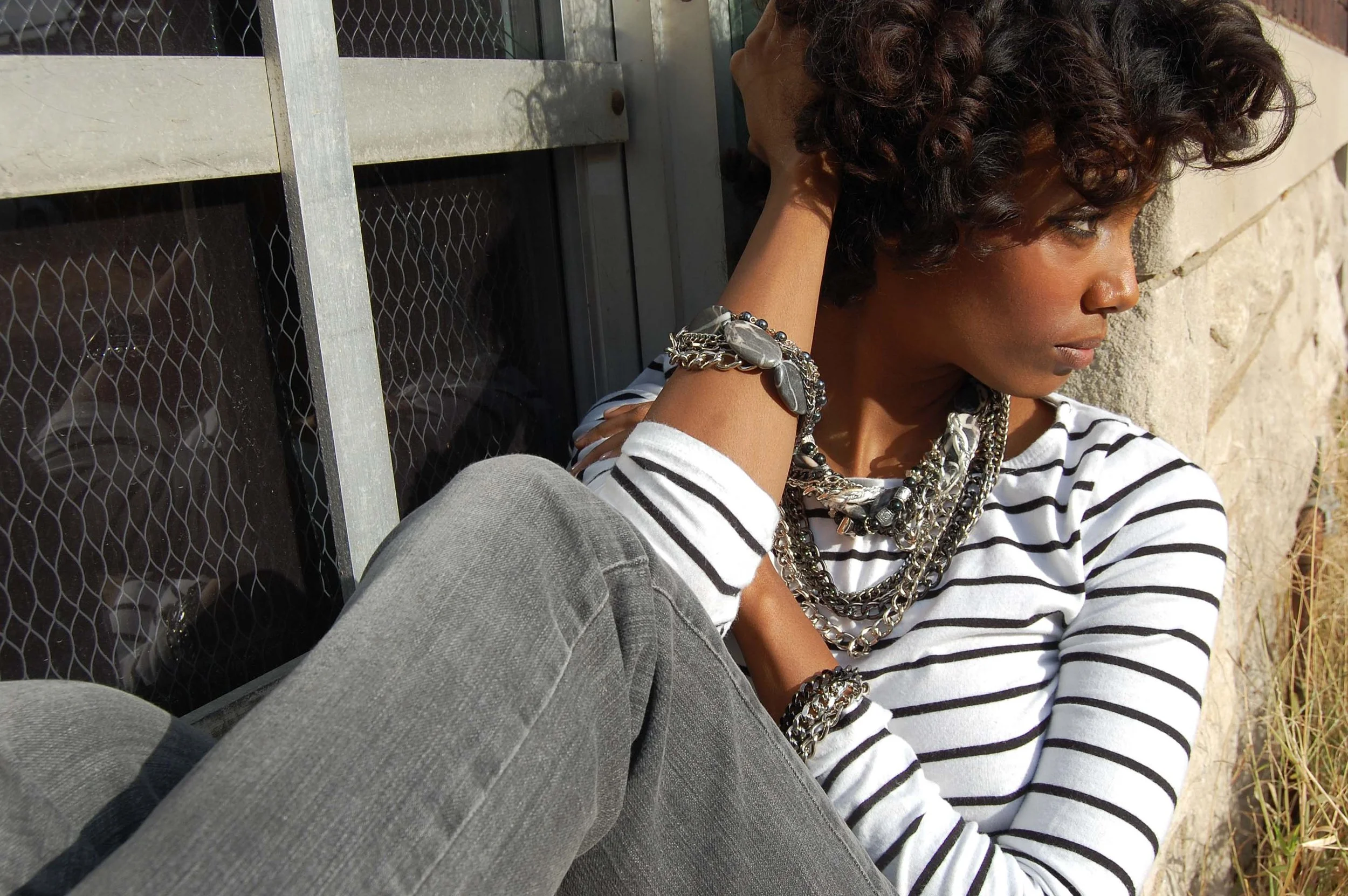 Woman with curly hair sitting against a window and stone wall, wearing a striped shirt and multiple silver jewelry layers.