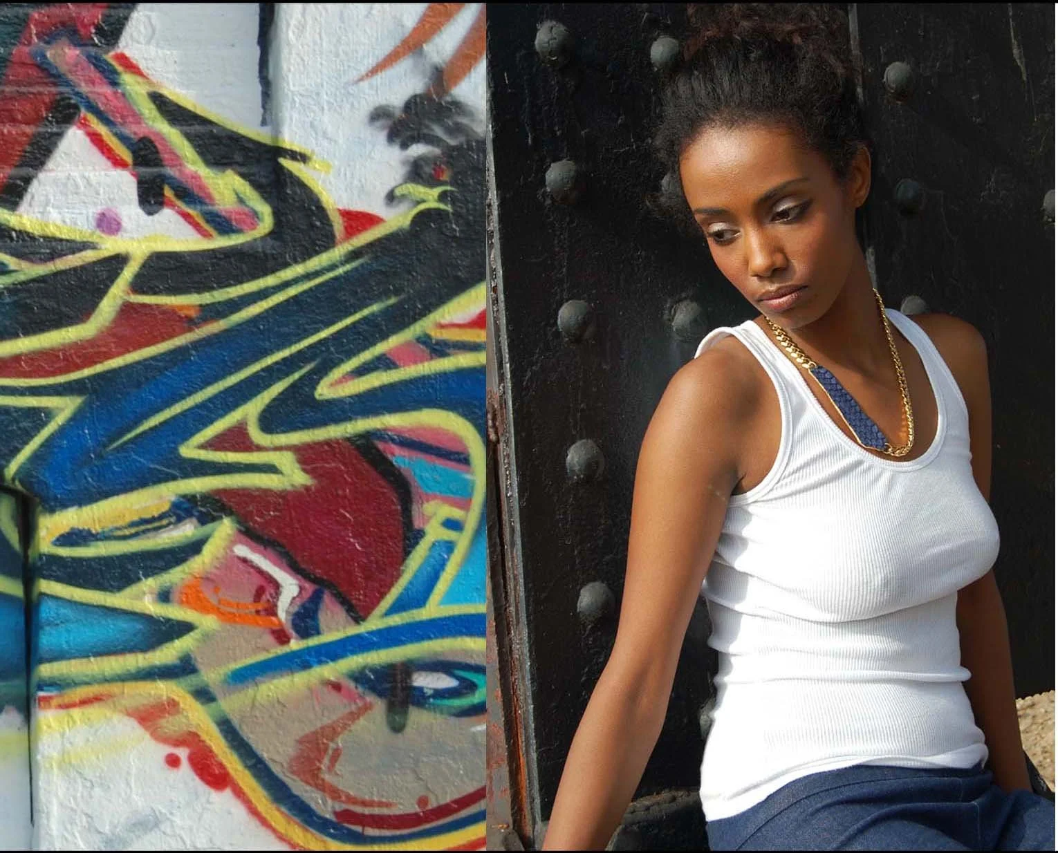 A woman with dark curly hair, wearing a white tank top and gold chain necklace, leaning against a black, studded metal door, looking down. The door has large bolts. Next to her, a colorful graffiti mural is partially visible.