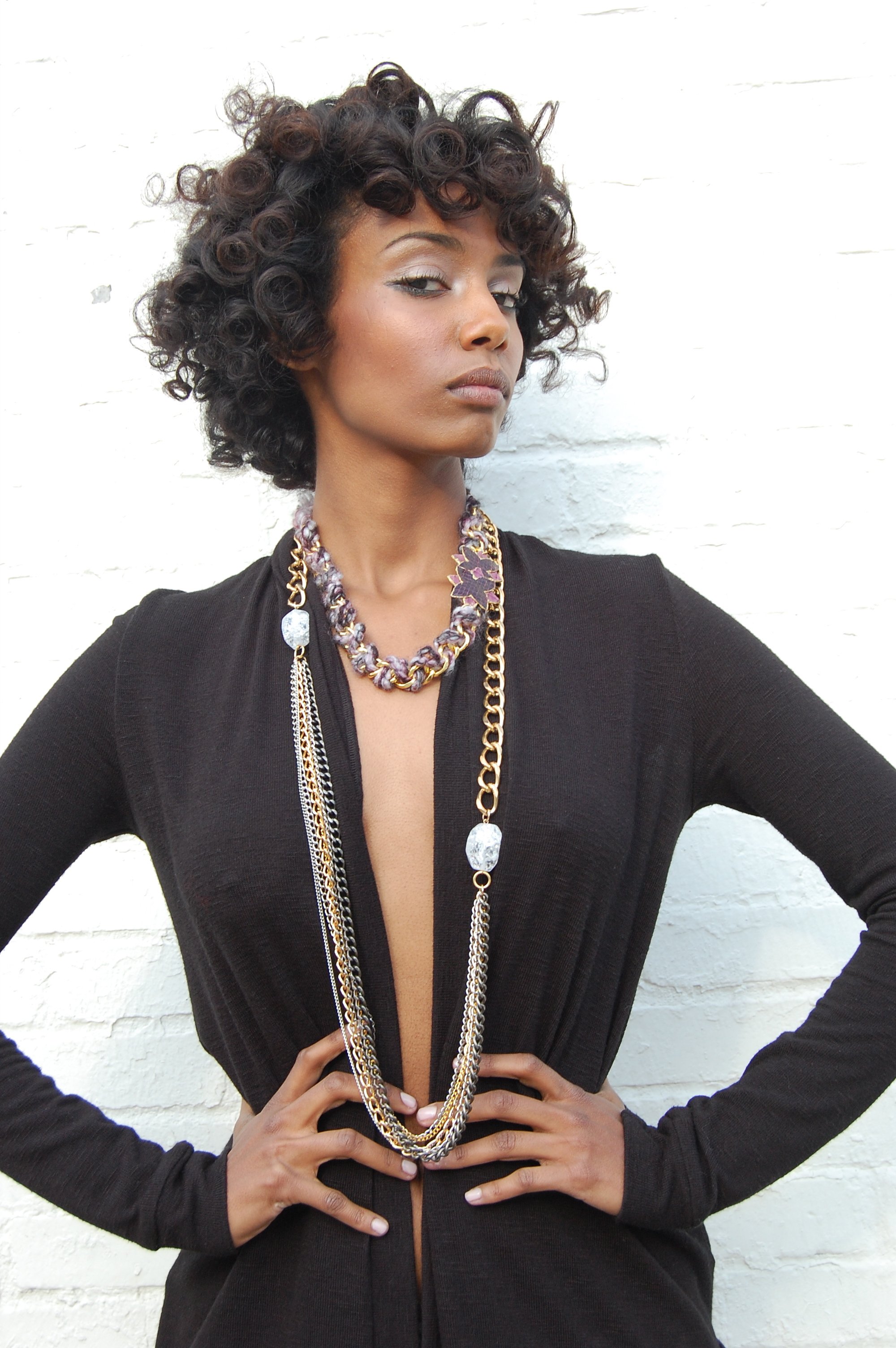 A woman with short, curly dark hair looking confidently at the camera, wearing a black outfit with multiple layered necklaces, poses against a white brick wall.