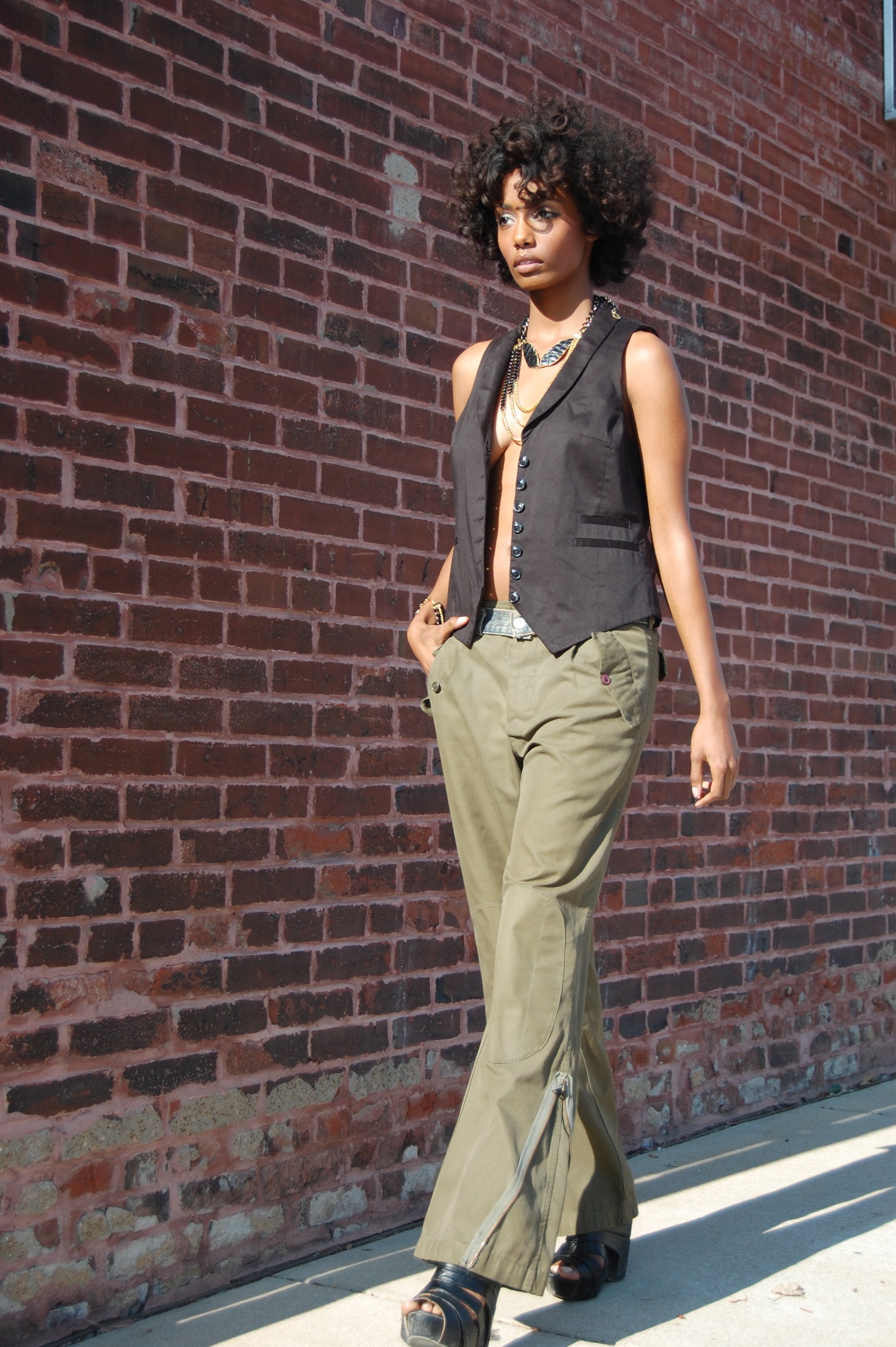 A woman with curly hair walking along a brick wall, wearing tan wide-leg pants with zippers, a sleeveless black vest, and black platform heels, accessorized with layered necklaces and bracelets.