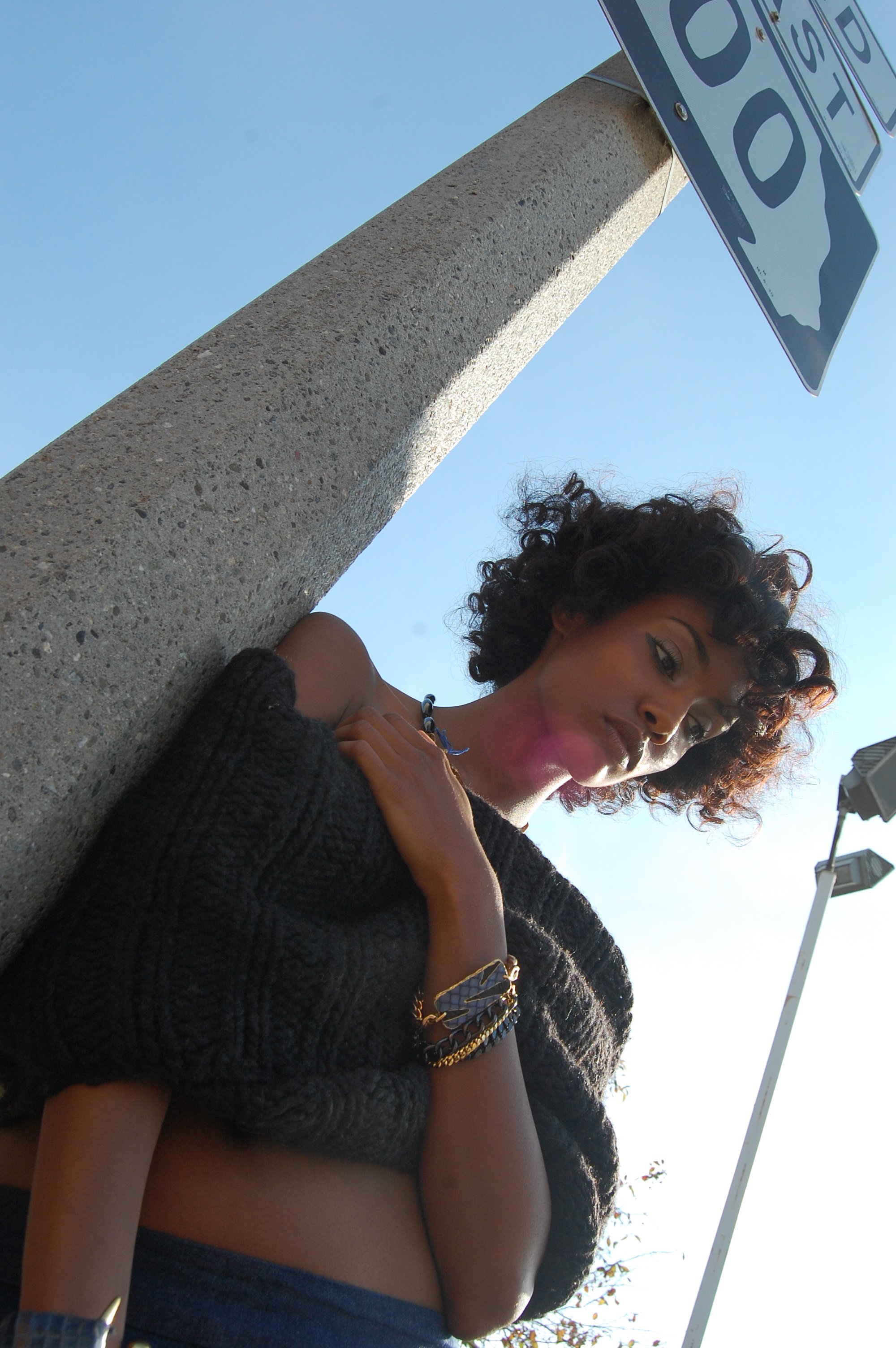 A young woman with curly hair standing near a concrete pole with a STOP sign above her. She is wearing a black sweater, jewelry, and is looking at the camera against a clear blue sky.