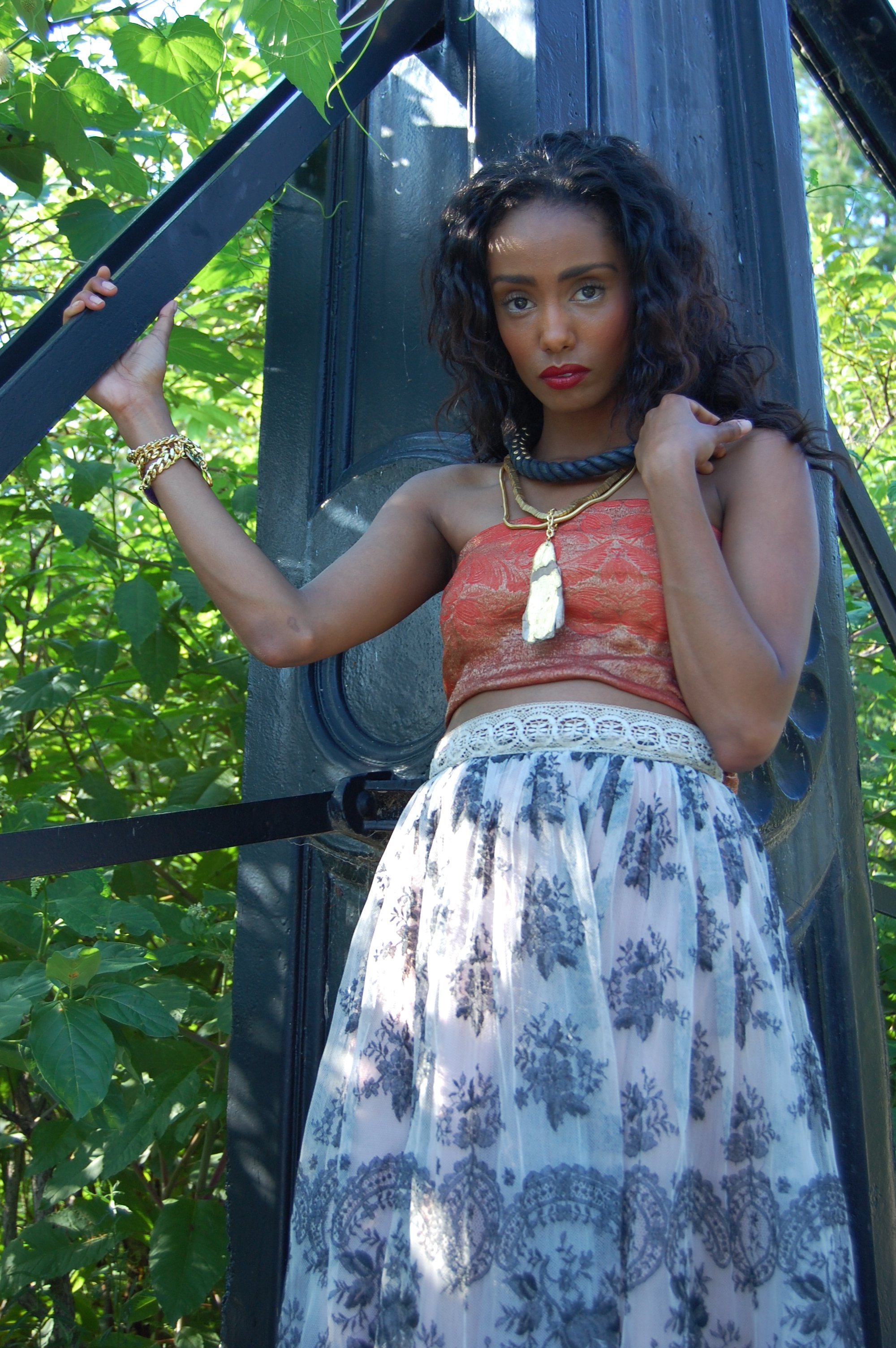 A woman with curly black hair wearing a red top, white patterned skirt, gold jewelry, and a black necklace with a feather pendant, standing outdoors next to a black metal structure surrounded by green foliage.