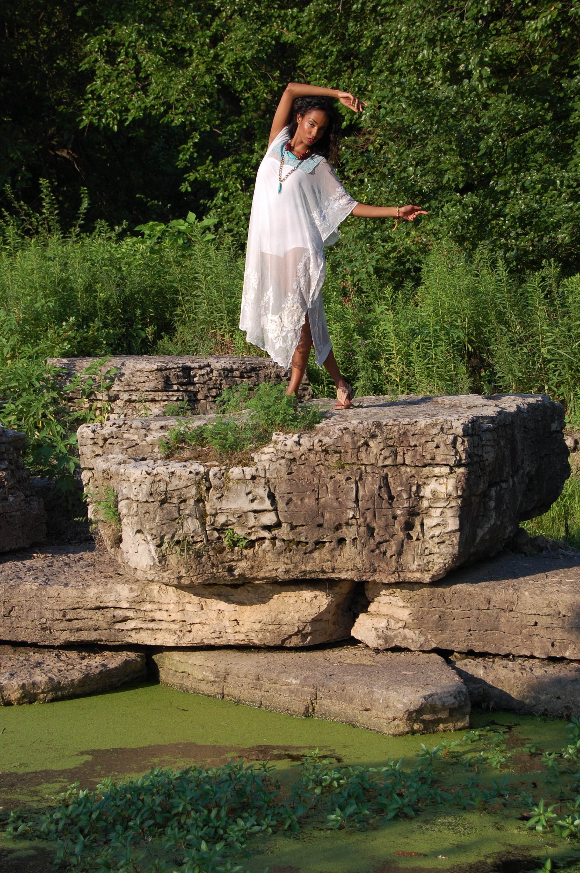 A woman standing on large rocks near a pond with green algae, surrounded by lush greenery, wearing a white dress and jewelry.