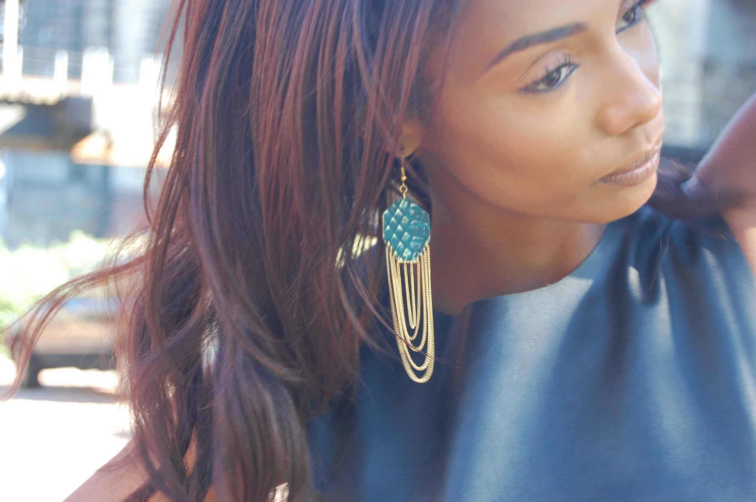 Close-up of a woman with long, wavy red hair wearing a blue top and large dangling turquoise and gold earrings, outdoors with blurred background.