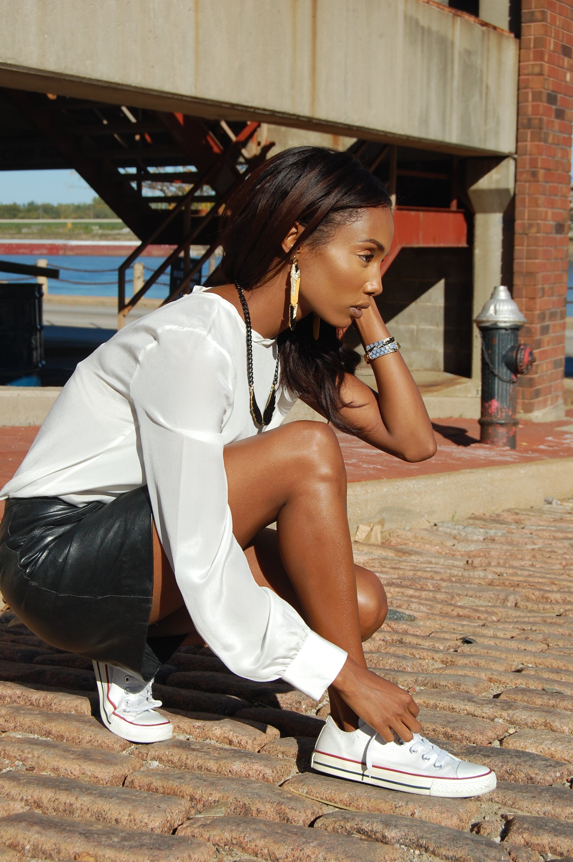 A young woman with dark hair and dark skin, wearing a white blouse, black leather shorts, and white sneakers, crouching on a brick sidewalk near a waterfront with a bridge and brick building in the background.