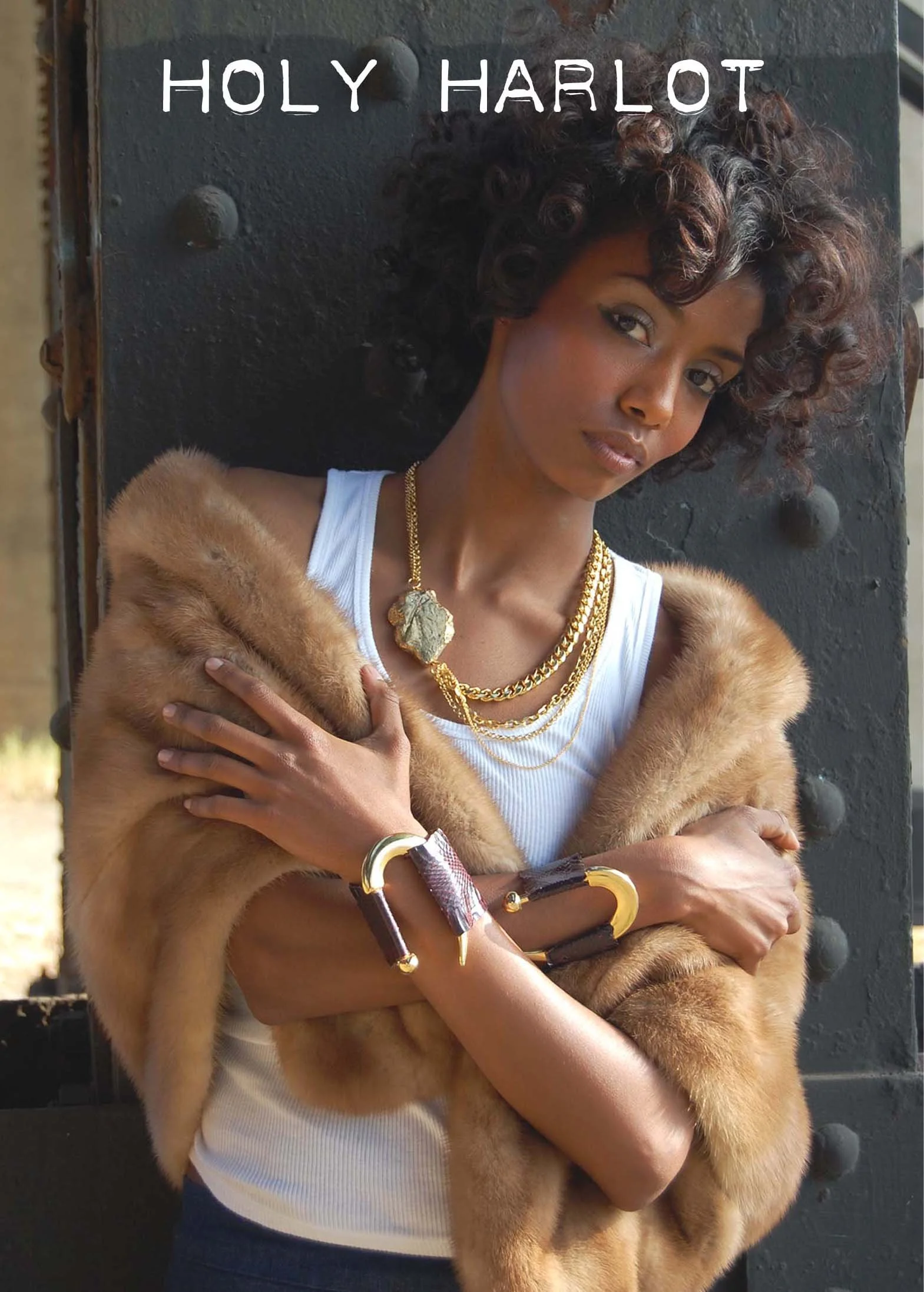 A woman with curly hair wearing a white tank top, gold jewelry, and a fur coat, standing against a large metal structure with the words 'HOLY HARLOT' at the top.