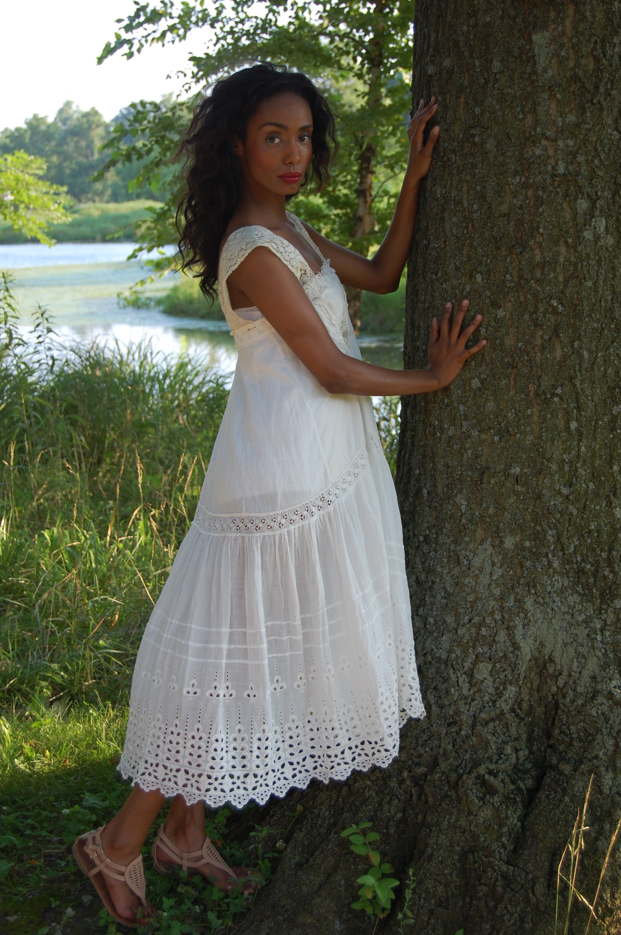 A woman in a white dress stands by a tree near a river, surrounded by greenery.