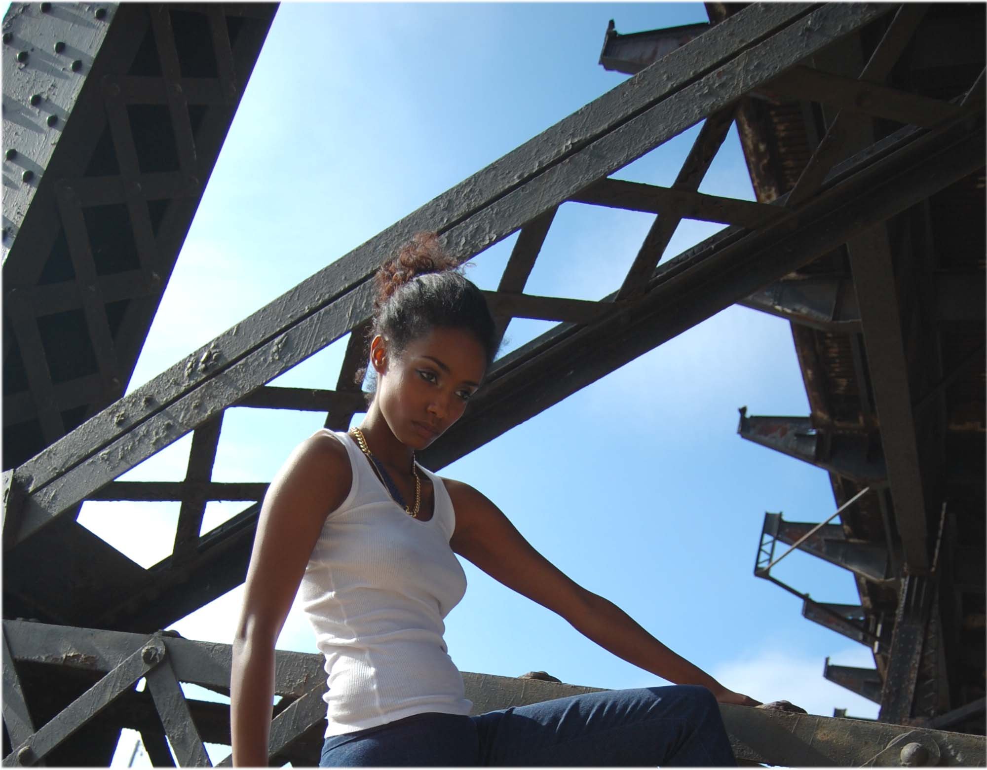 A young woman with dark skin and braided hair tied up in a bun, wearing a white sleeveless shirt and dark pants, sitting on a metal structure under blue sky with industrial buildings around.