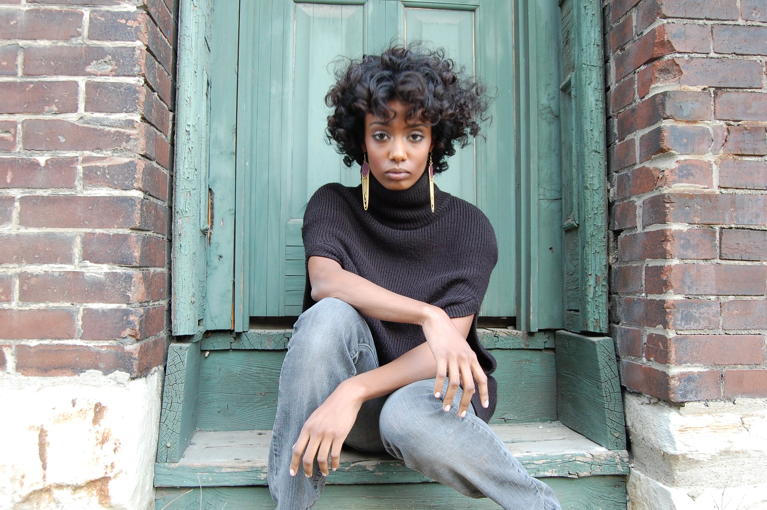 A young woman with curly hair wearing a black turtleneck sweater and gray jeans sitting on a step outside in front of a turquoise wooden door.
