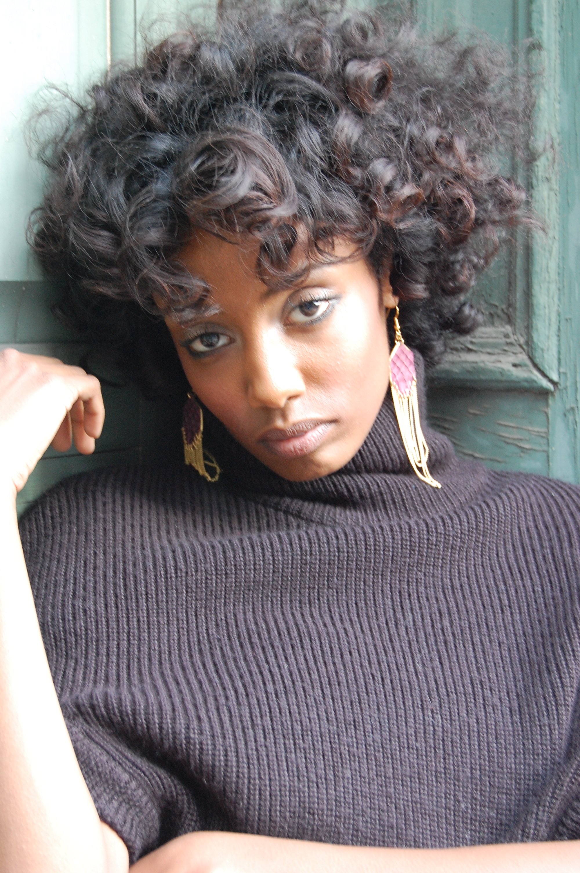 A woman with curly hair wearing a black turtleneck and gold and pink earrings standing against a green wooden wall.
