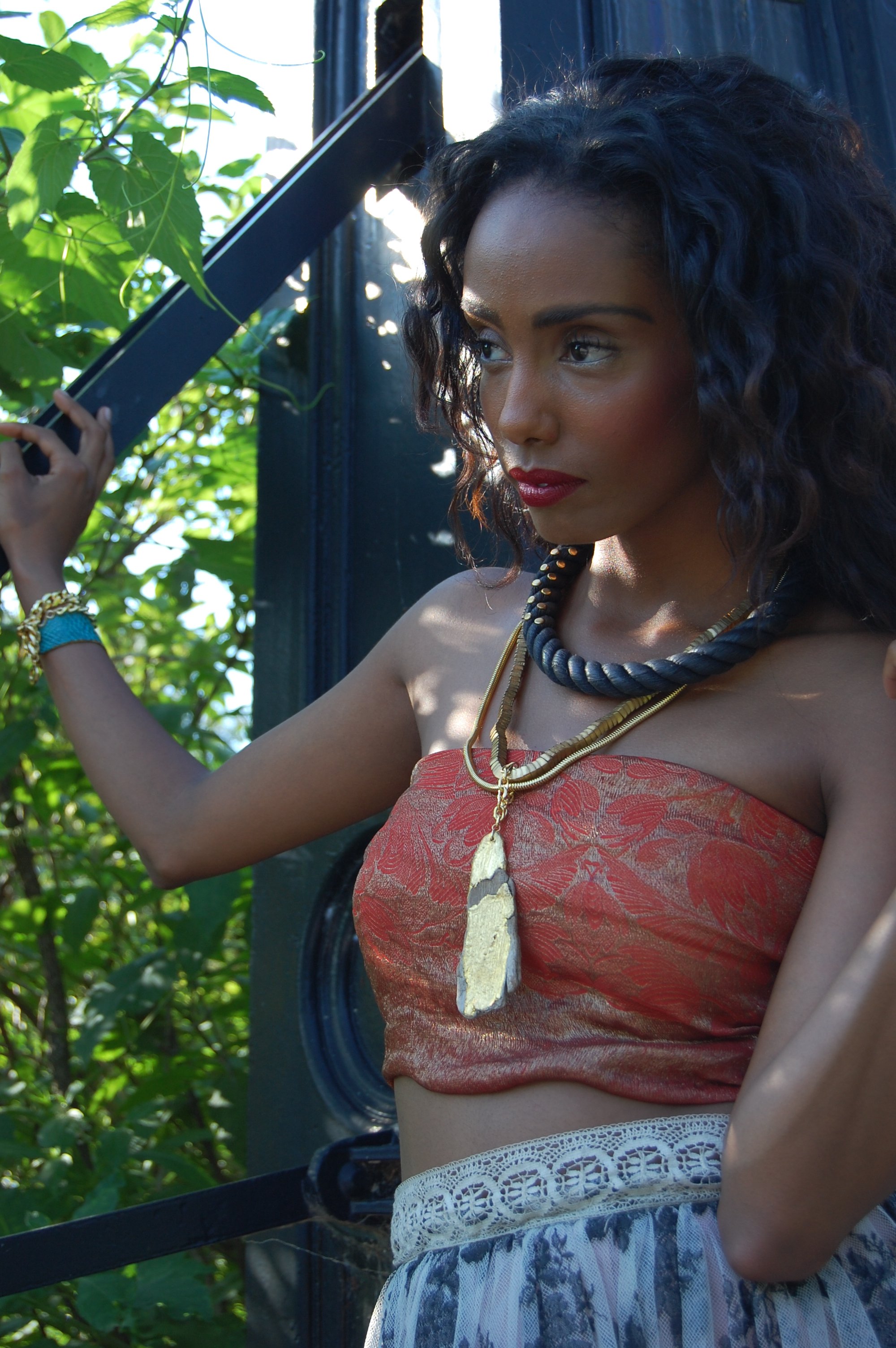 A woman with dark curly hair wearing a strapless red top with gold floral patterns, layered necklaces, and a large stone pendant necklace. She is outdoors, near a green leafy background, holding onto a metal structure with her left arm.