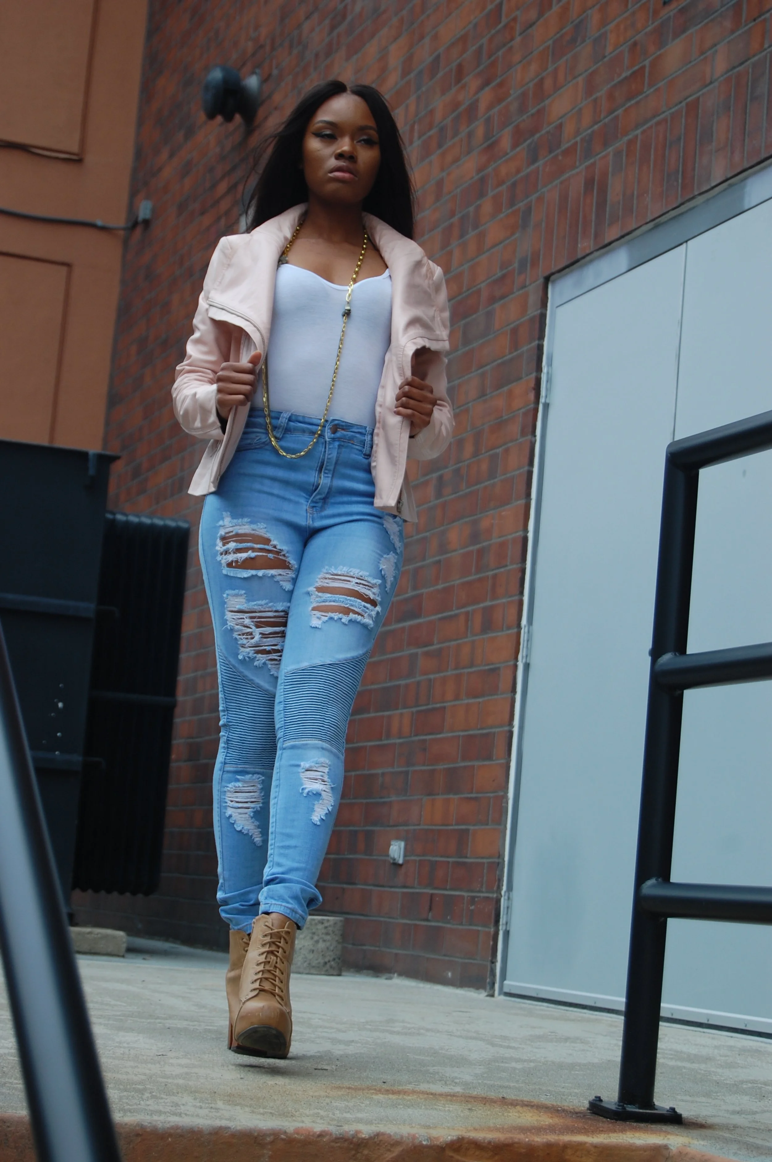 Young woman with long black hair walking outdoors against a brick wall, wearing a light pink jacket, white top, ripped blue jeans, tan high-heeled boots, and gold jewelry.