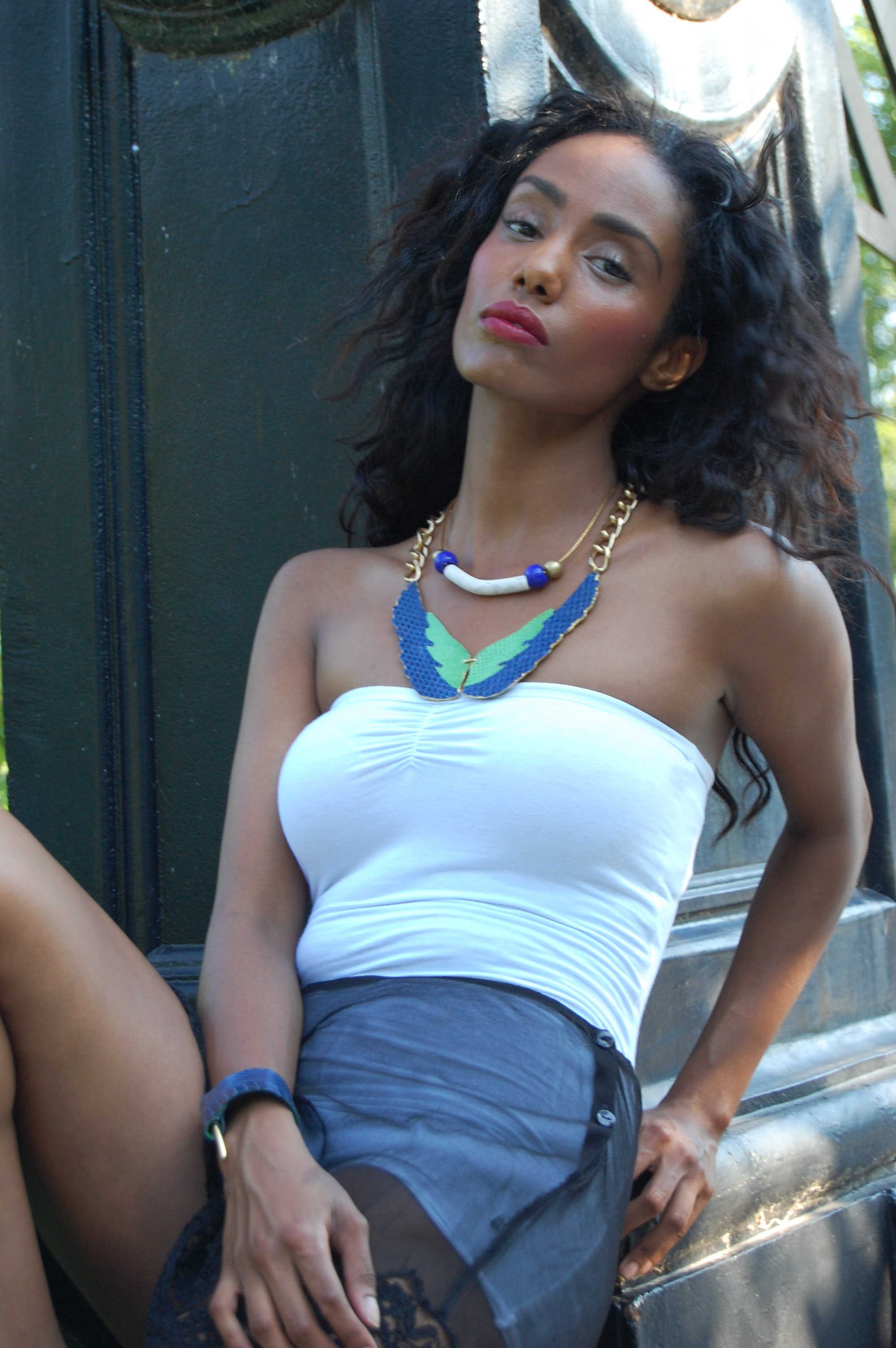 A woman with long curly hair sitting outdoors, wearing a white strapless top, a dark skirt, and colorful necklaces.