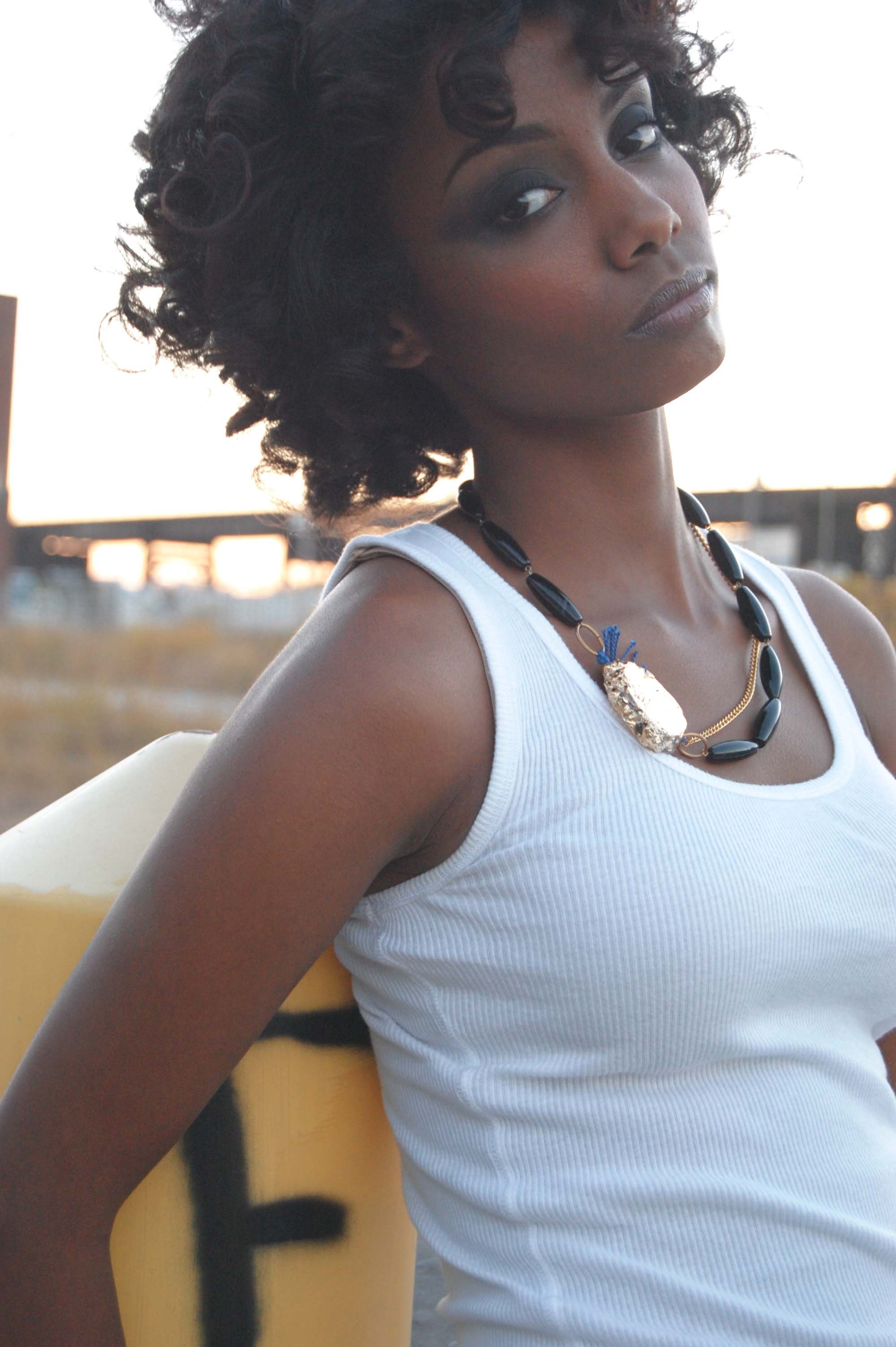 Portrait of a young woman with curly hair, wearing a white tank top and a black beaded necklace, standing outdoors at sunset with a blurred urban background.