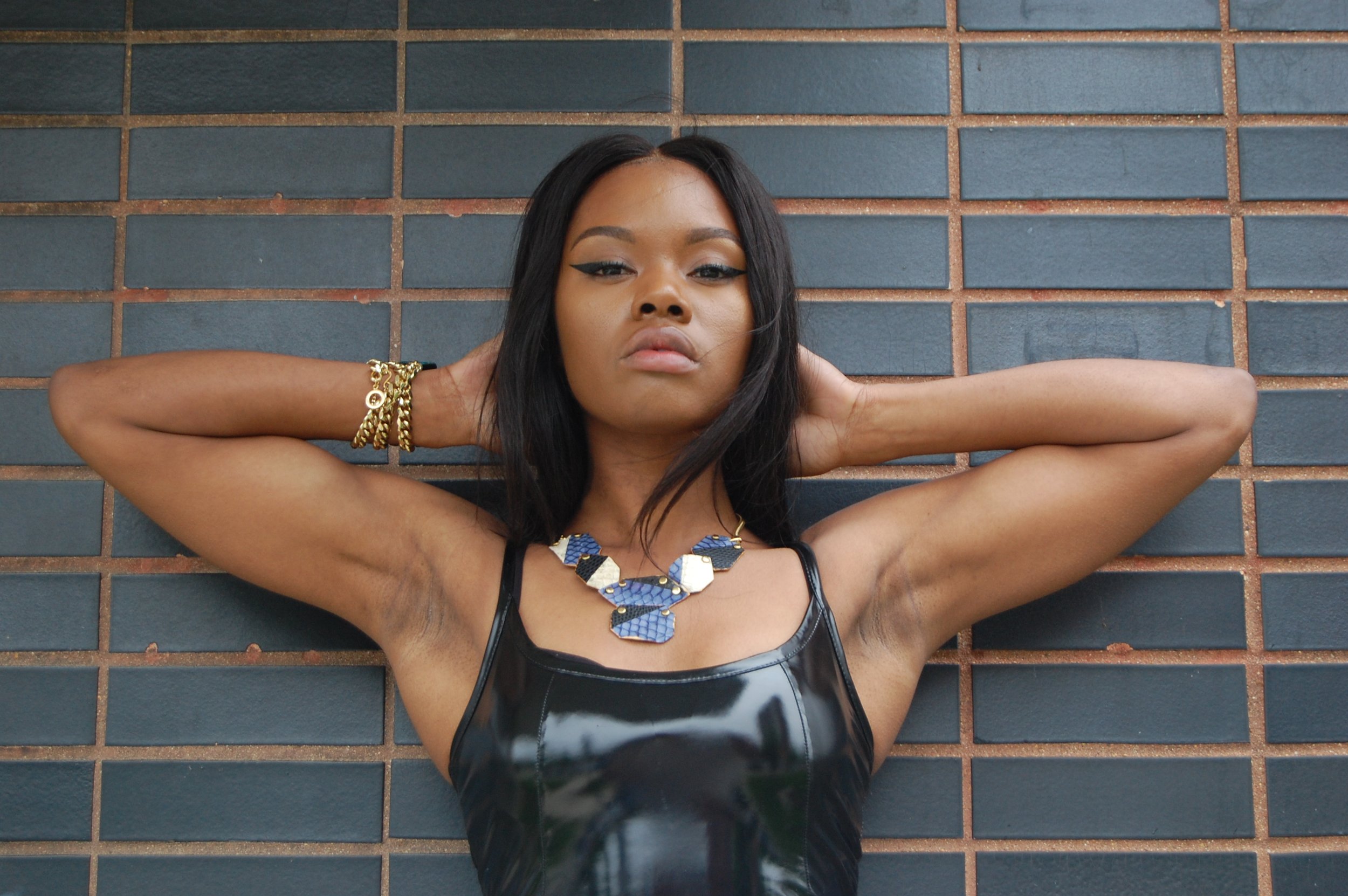 A woman with dark hair and makeup, wearing a leather top, gold bracelets, and a blue and white necklace, standing against a brick wall with her hands behind her head.Holy Harlot Jewelry photographed by artist Magda Parasidis, fashion photographer.