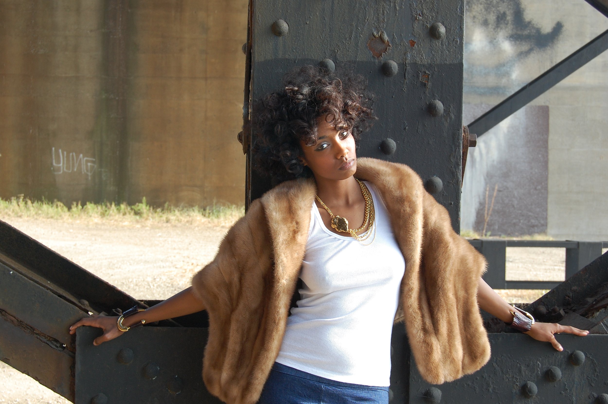 A woman with curly hair wearing a brown fur coat, white top, and jewelry, posing outdoors against a metal structure. Holy Harlot Jewelry photographed by Magda Parasidis.