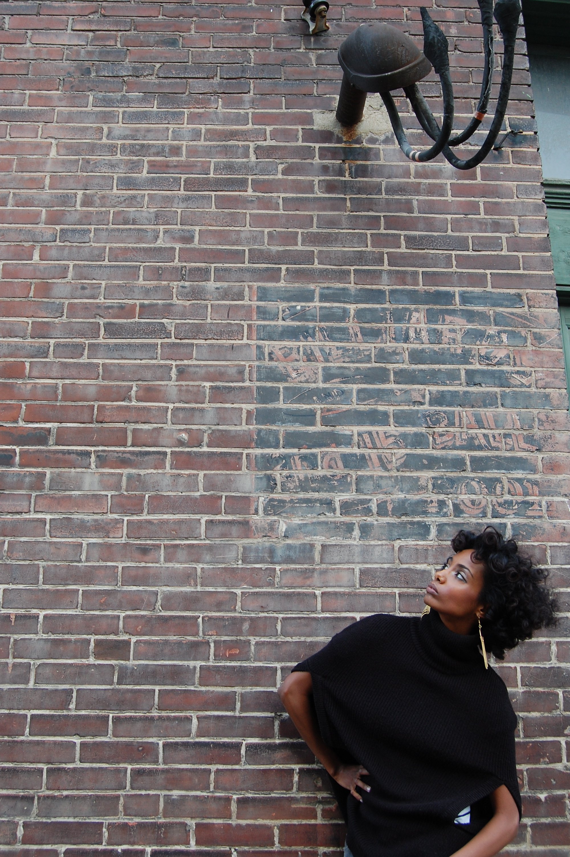 A woman with curly hair wearing a black sweater with sleeves rolled up, standing against a brick wall with faded graffiti, looking to her right with a serious expression.