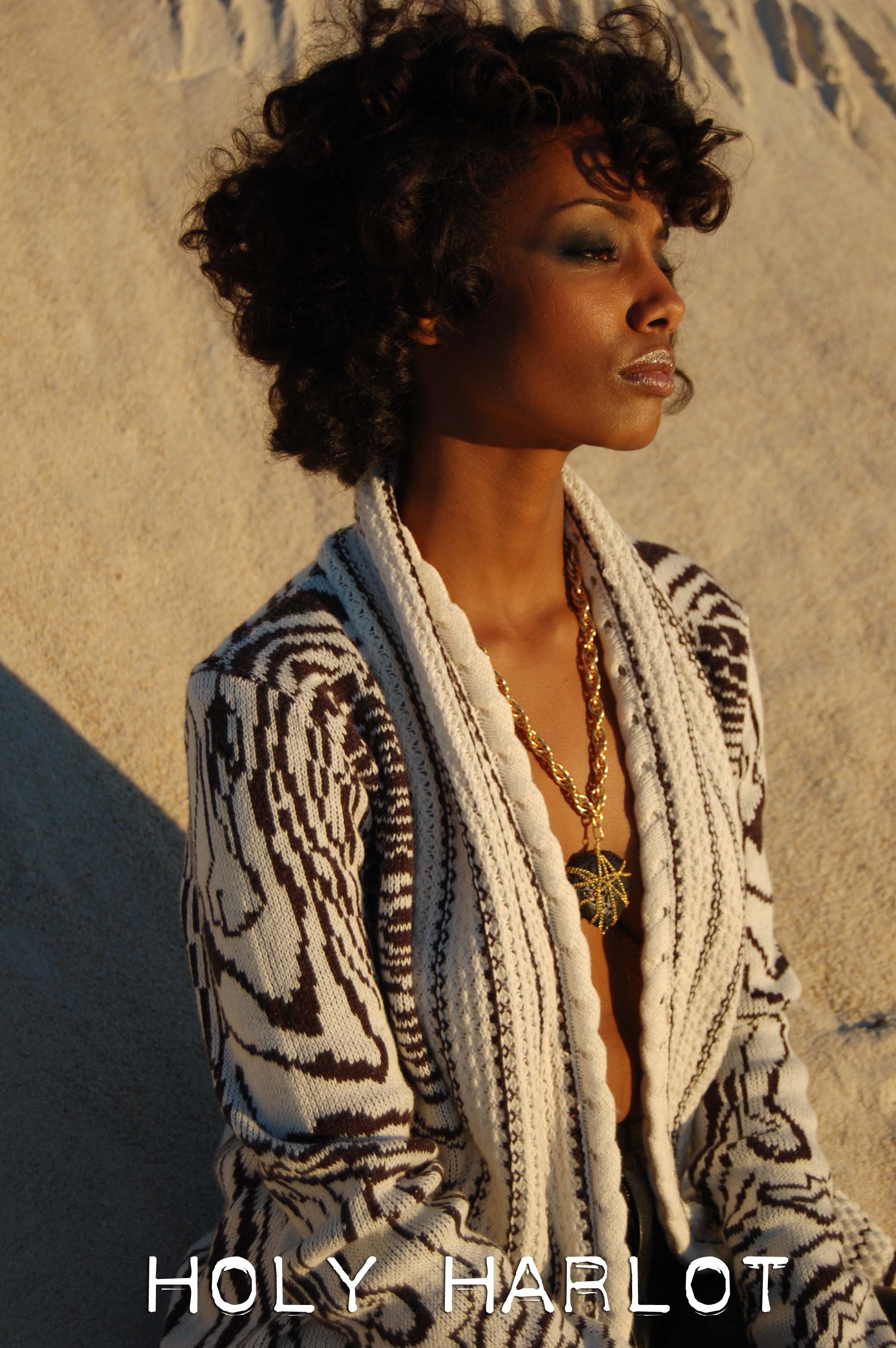 A woman with dark skin tone is standing against a textured, beige wall with her eyes closed and head slightly tilted. She is wearing a patterned sweater and a gold necklace with a heart pendant by Holy Harlot Jewelry photographed by Magda Parasidis.