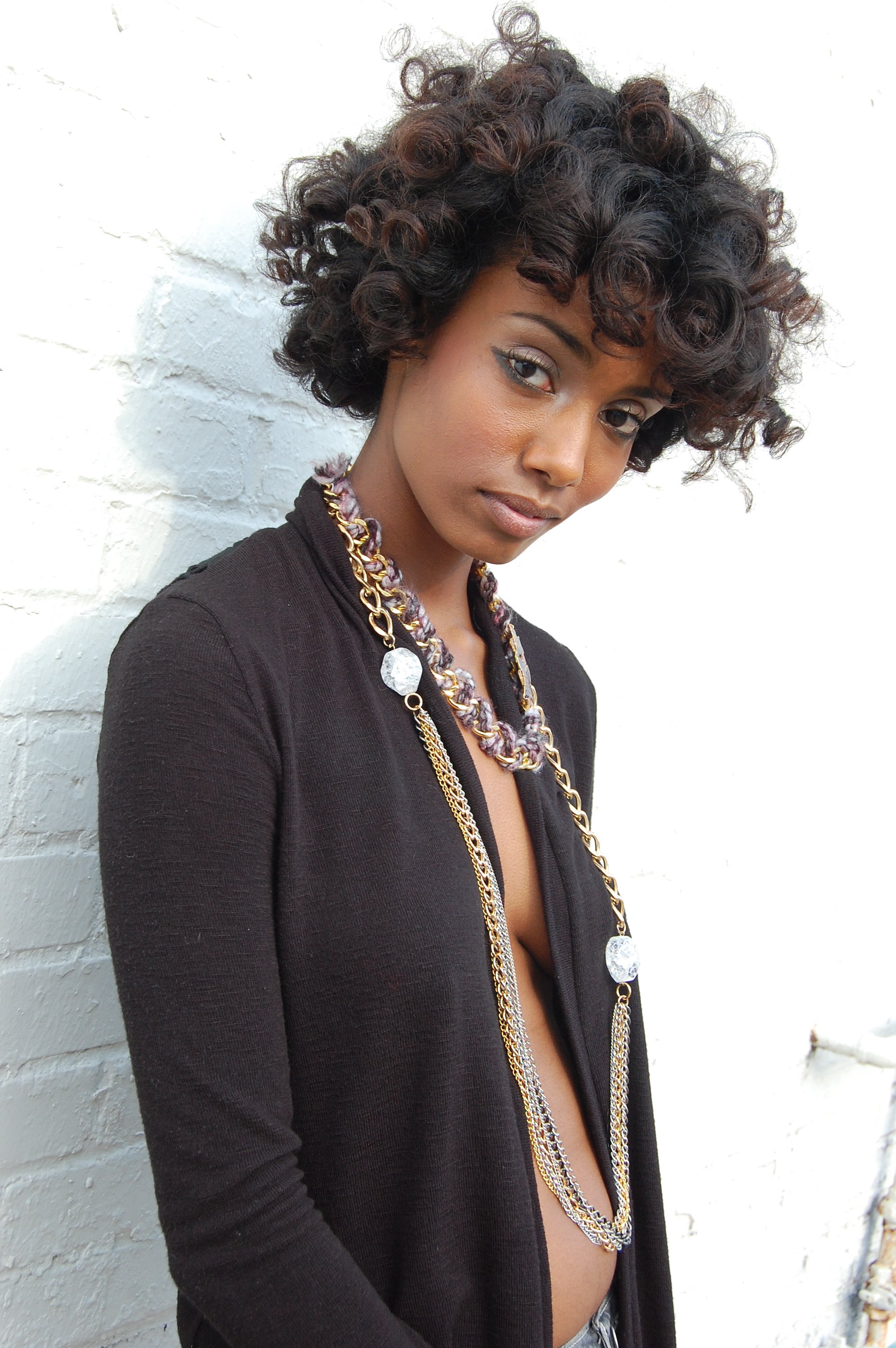 Fashionably dressed woman with curly hair and layered necklaces standing against a white brick wall.