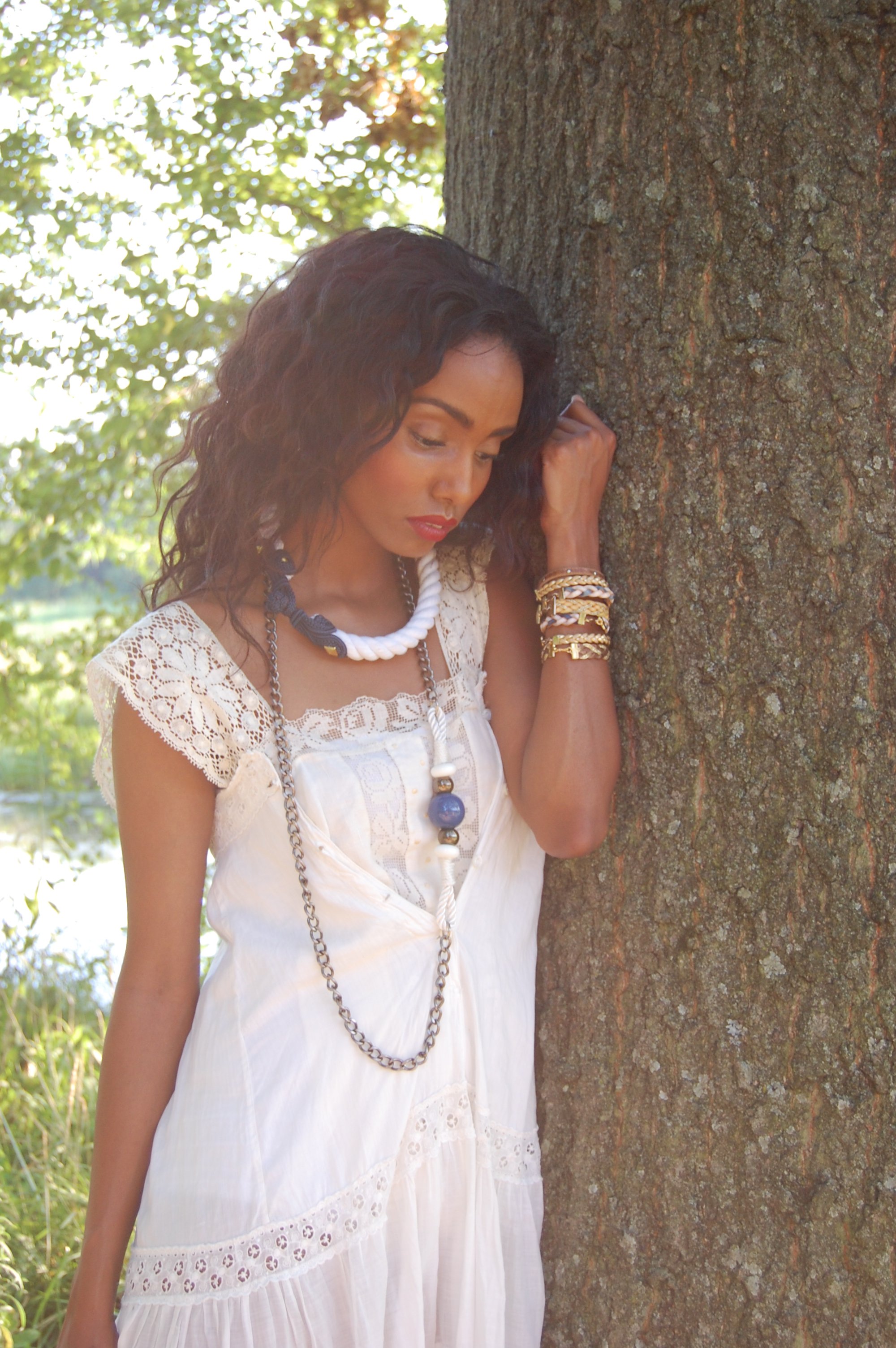 A woman with dark curly hair wearing a white lace dress, multiple necklaces, and bracelets, leaning against a tree in a natural outdoor setting with sunlight filtering through the leaves.