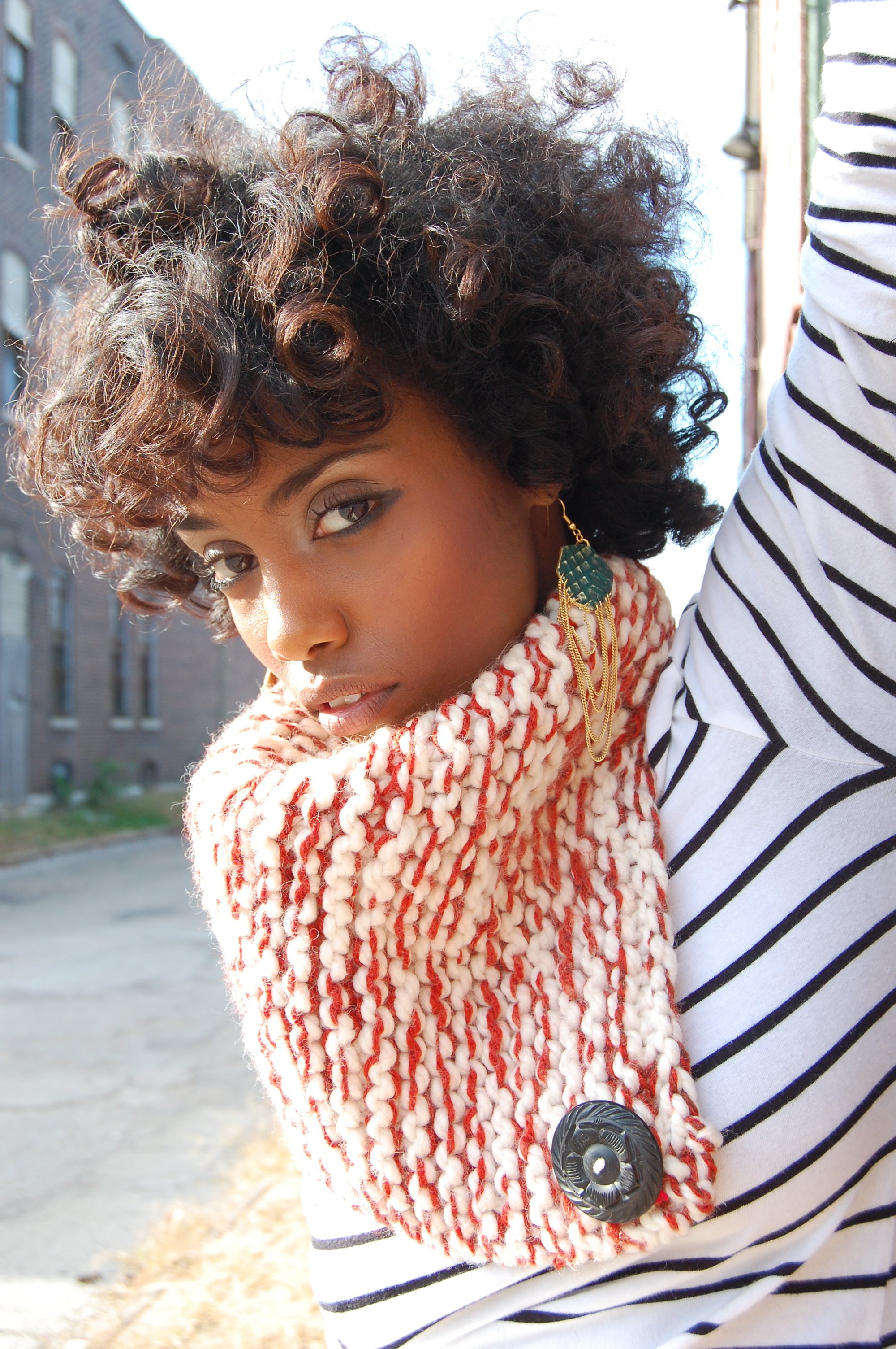 A woman with curly hair wearing a striped shirt and a chunky knit scarf, outdoors on a sunny day.