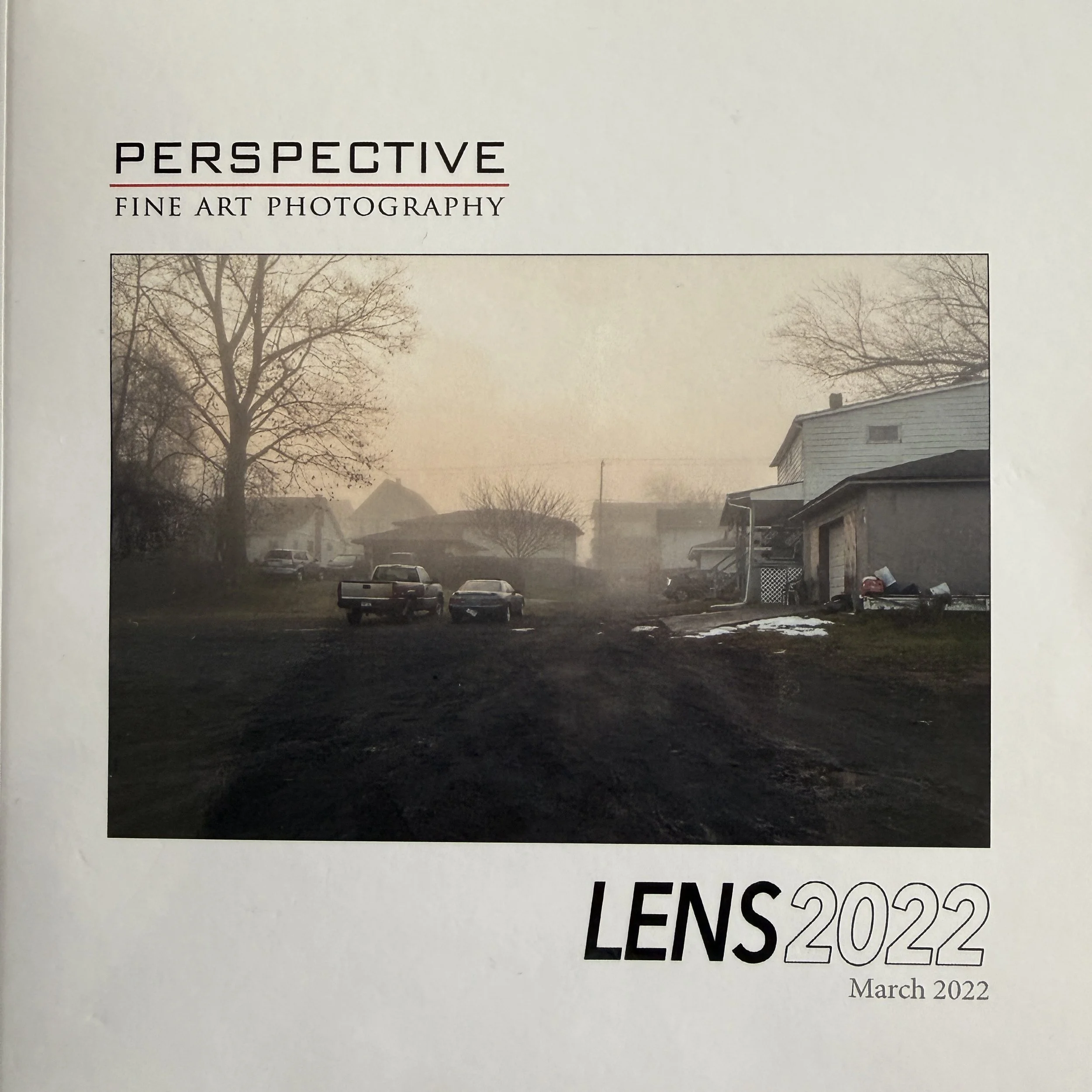 Cover of Lens 2022 catalogue from Perspective Gallery, featuring Magda Parasidis' photocollage The Block.  A misty neighborhood scene with several vehicles parked on unpaved, muddy ground, trees without leaves, and houses  on a foggy day.
