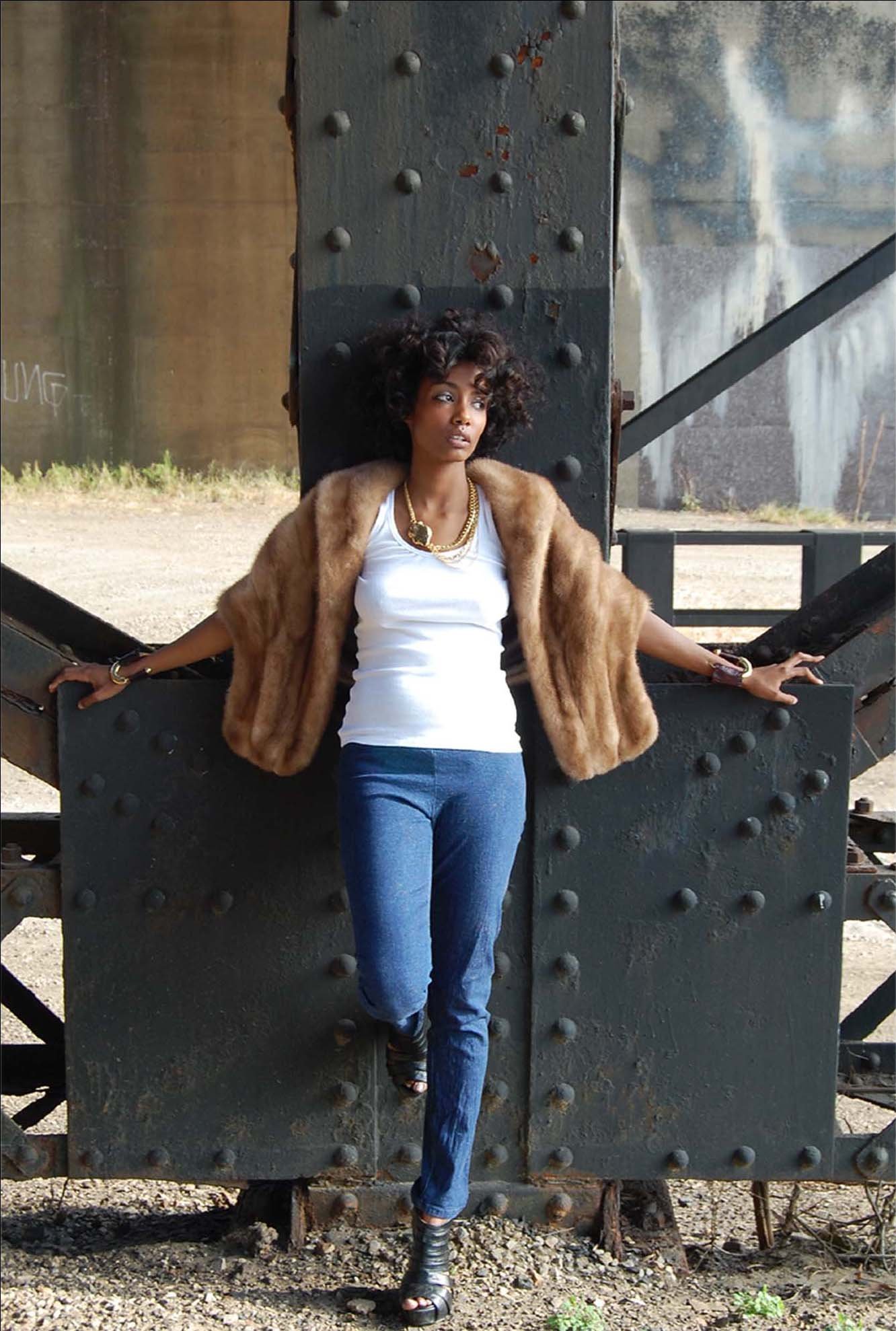 A woman stands in front of a large metal industrial structure outdoors. She wears a white top, blue jeans, black open-toed shoes, and a brown fur coat. She has layered gold necklaces and bracelets, with short, curly hair.